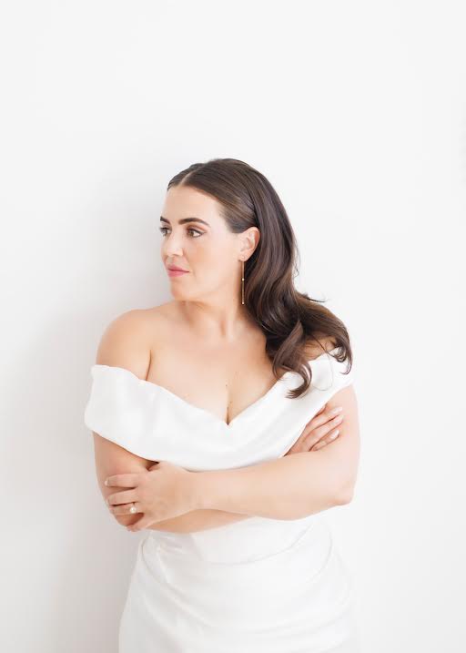 Woman with dark hair in a white off-shoulder dress, arms crossed, looking to the side, against a plain white background.