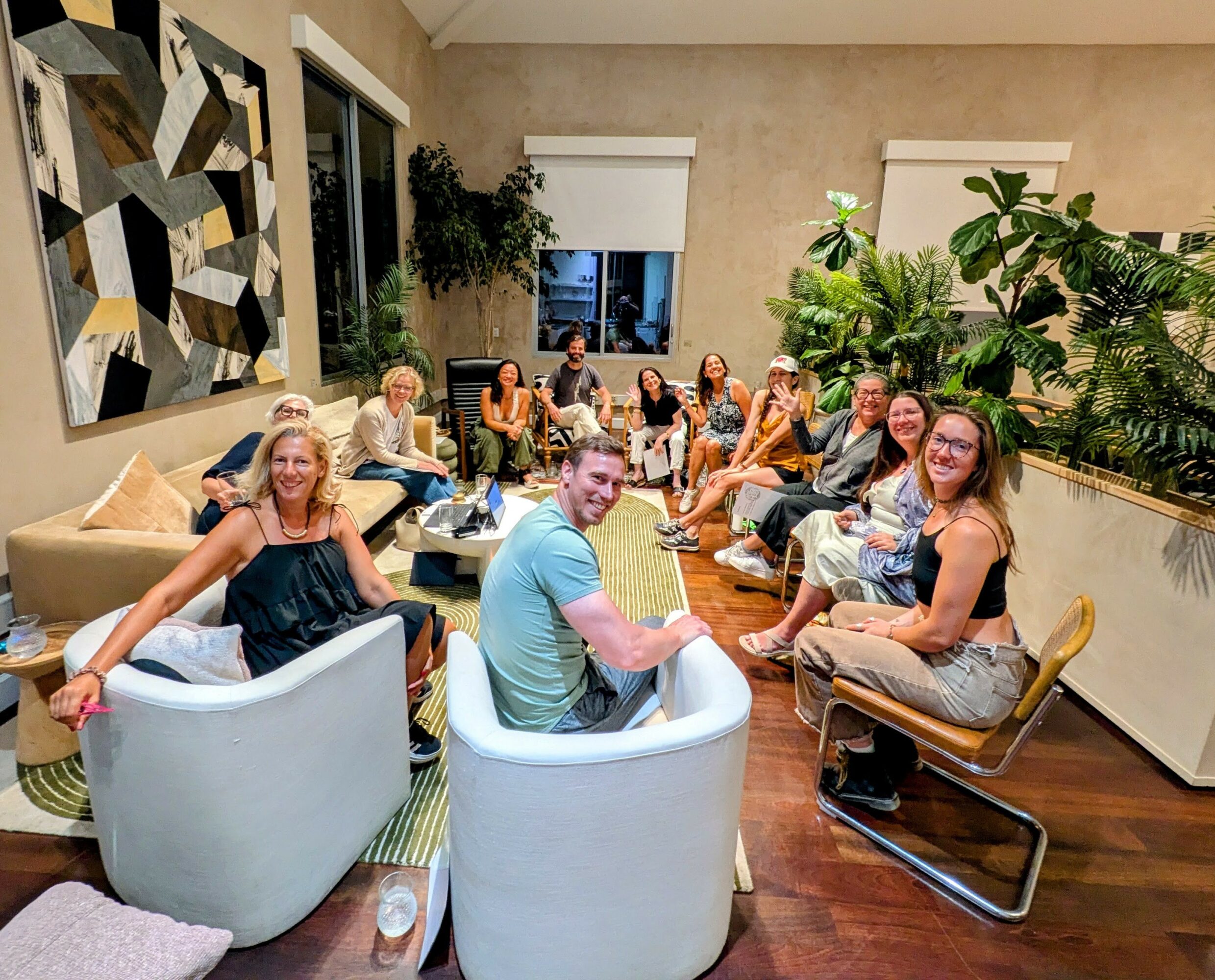 Group of people sitting and chatting in a living room with large windows, plants, and modern decor.