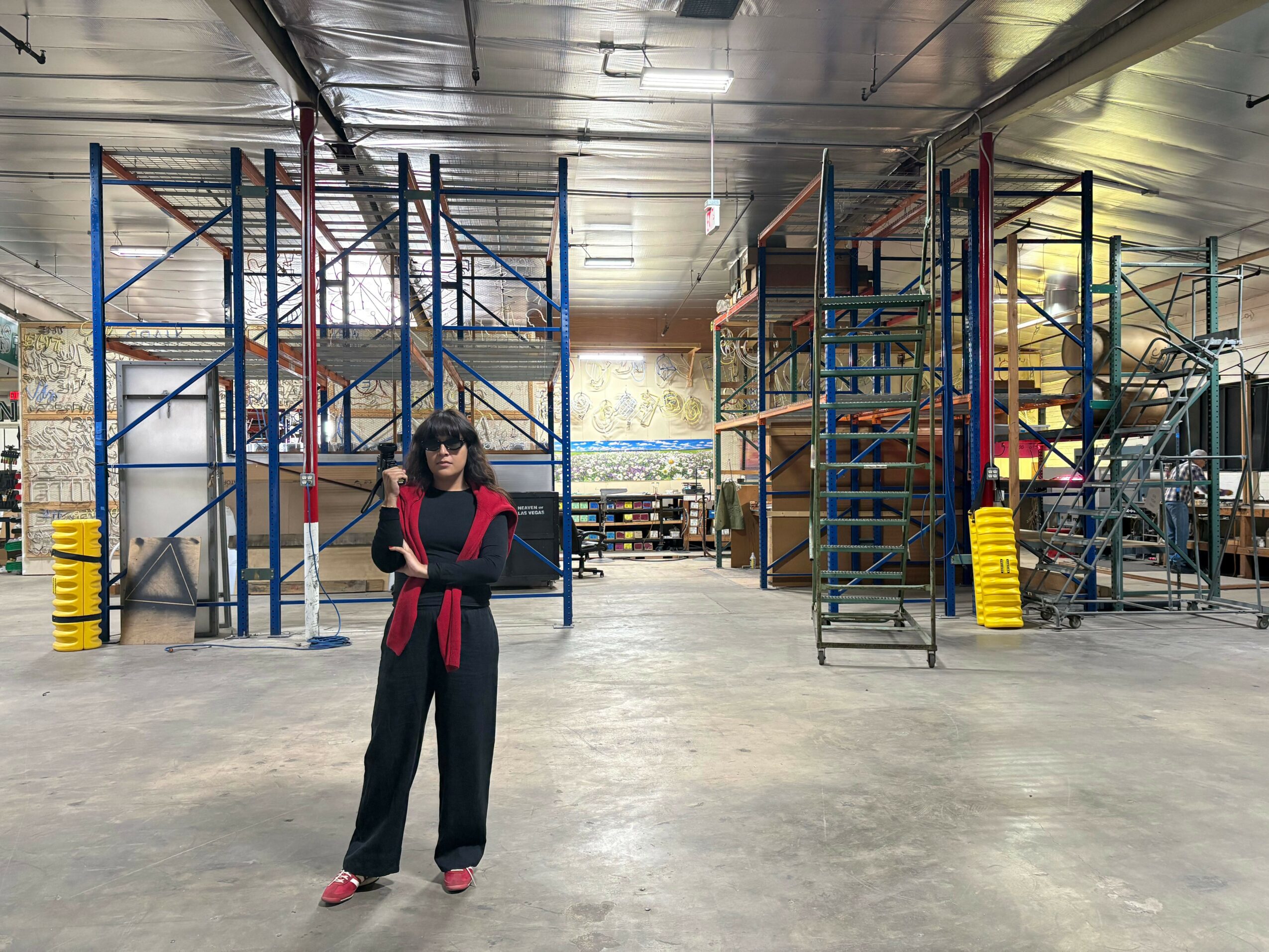 Person standing in an industrial warehouse with metal scaffolding and equipment, wearing dark clothing and sunglasses.