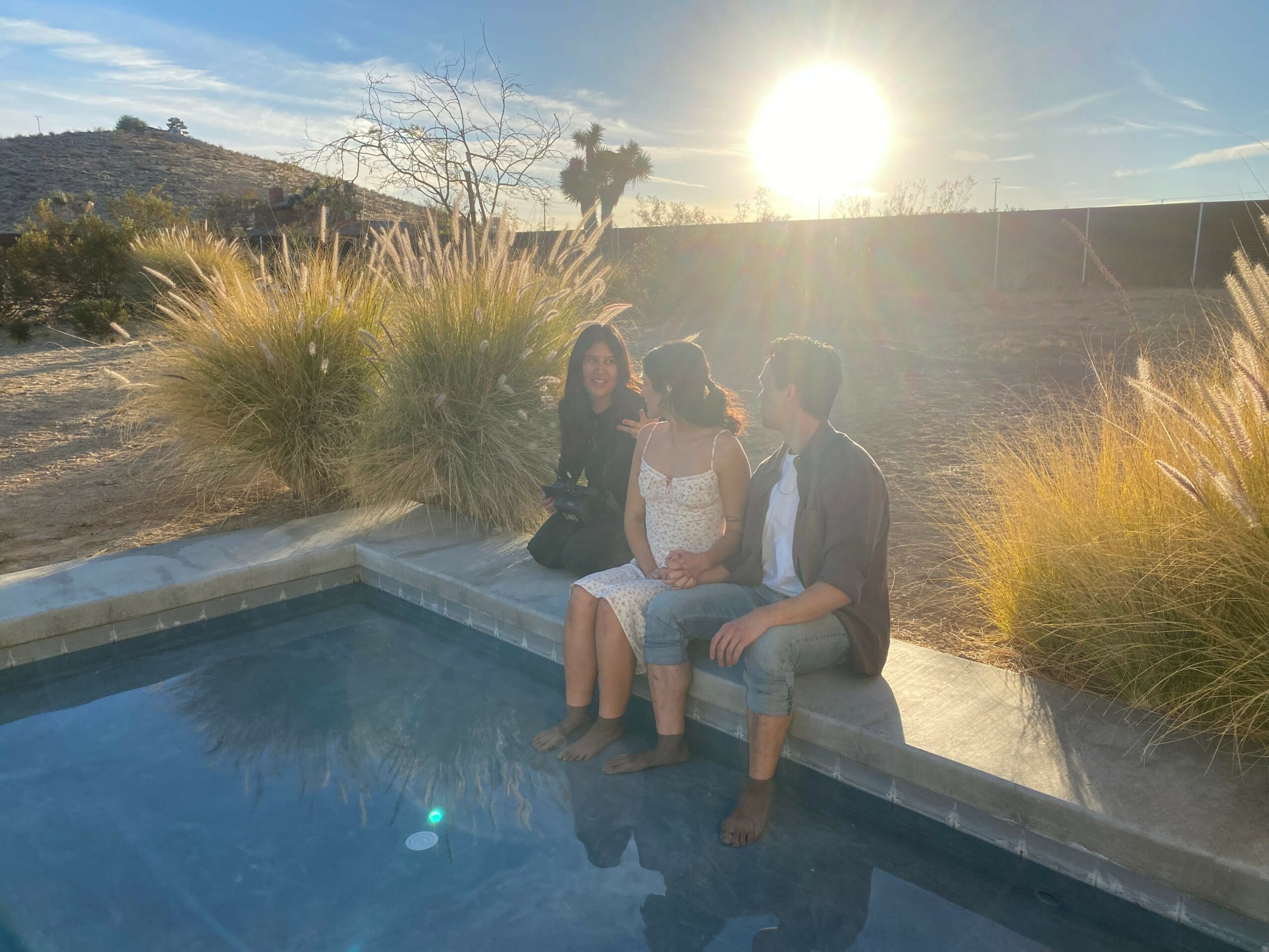 Three people sit by a pool on a sunny day, with a landscape and bright sun in the background.