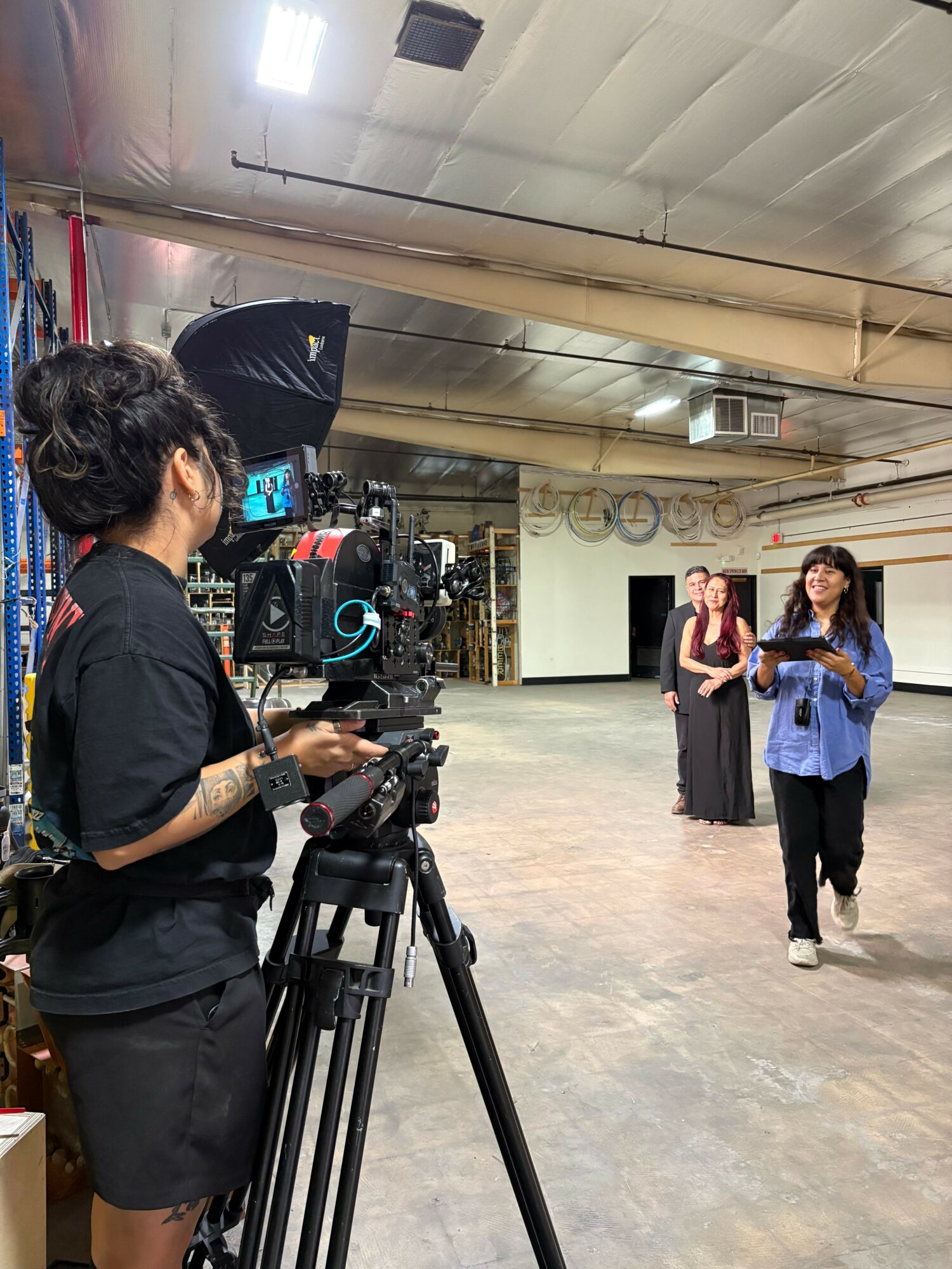 A woman operating a camera records two women standing in a spacious indoor area with a high ceiling.