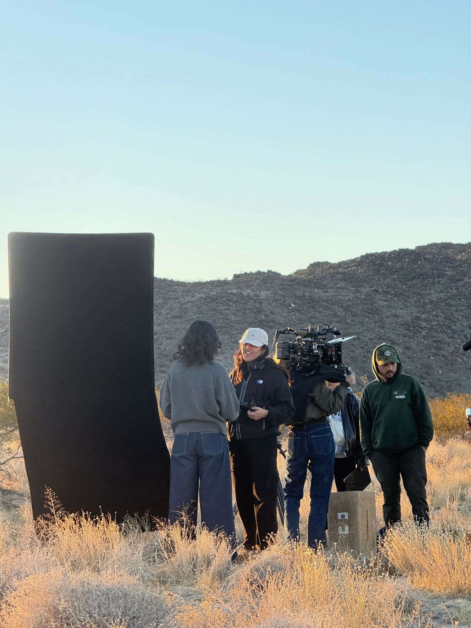 Group of people and camera crew outdoors near a large black structure, with a hill and clear sky in background.
