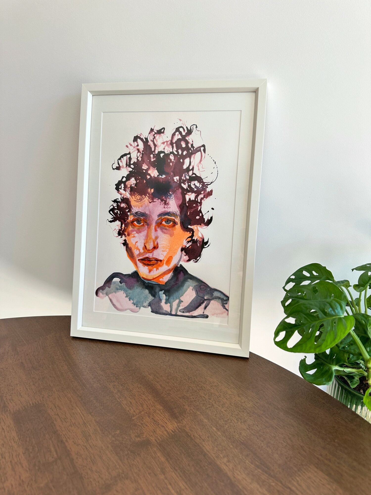 Framed watercolor portrait of a woman with curly hair, leaning against a white wall, next to a green plant.