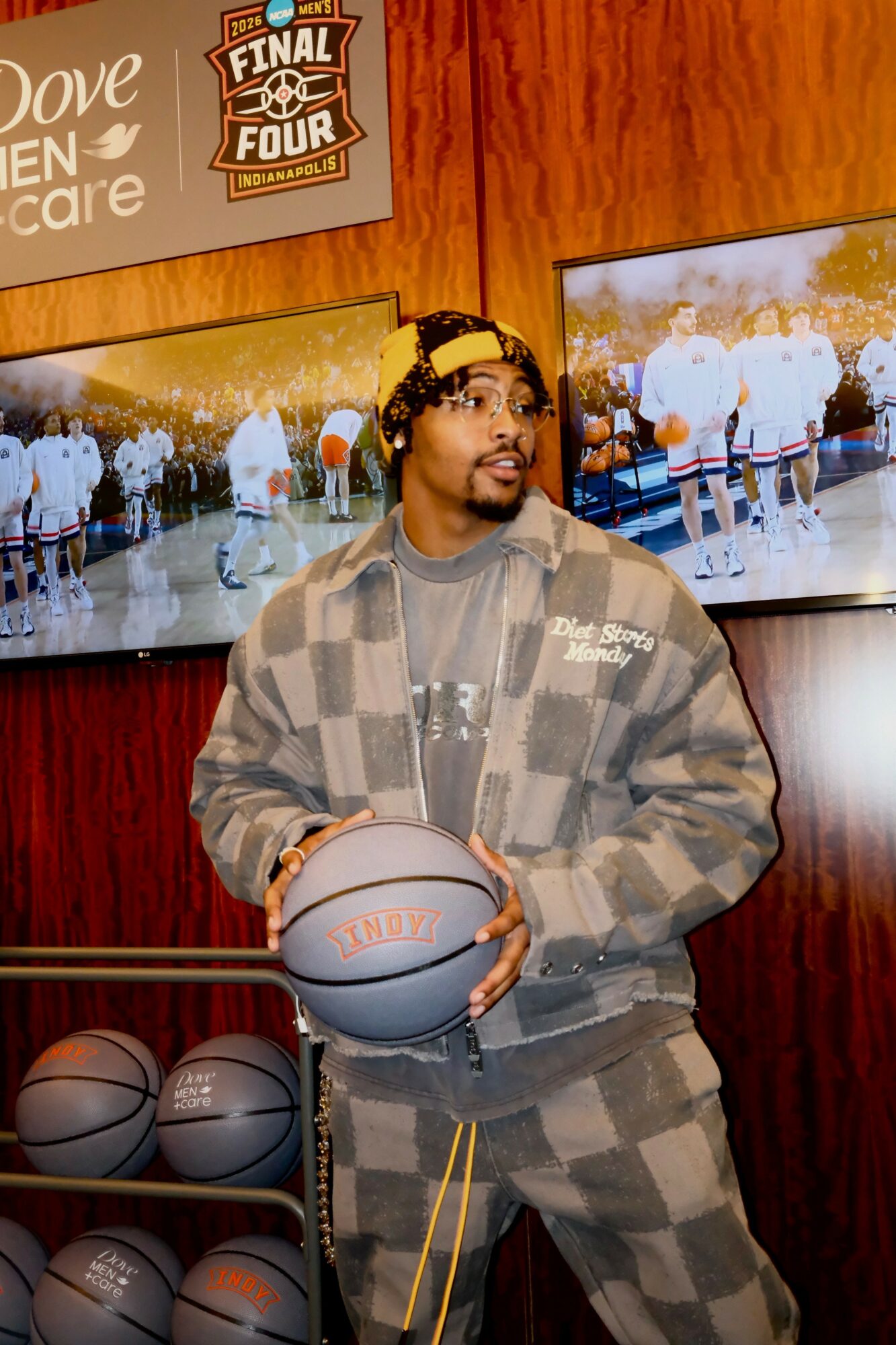 Man holding a basketball in a room with basketballs and TV screens, wooden wall background, wearing a checkered jacket and headband.
