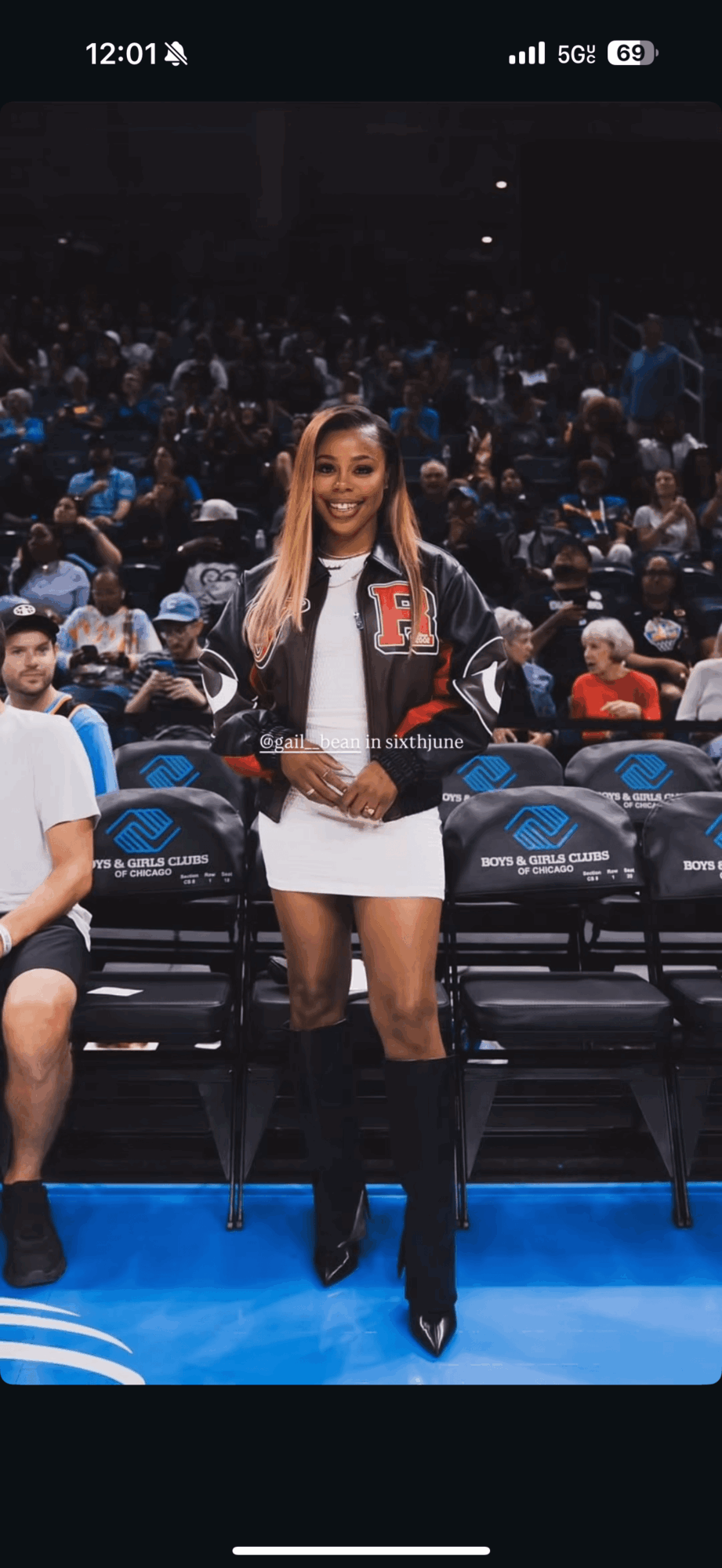 Young woman standing in sports arena, smiling, holding a paper, wearing a jacket and white dress, with audience in background.