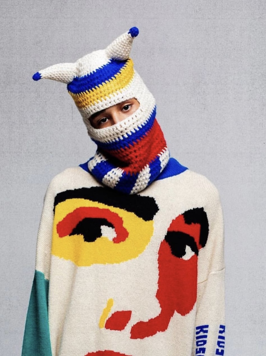 Person wearing a colorful knitted hat and sweater with face designs, against a plain background.