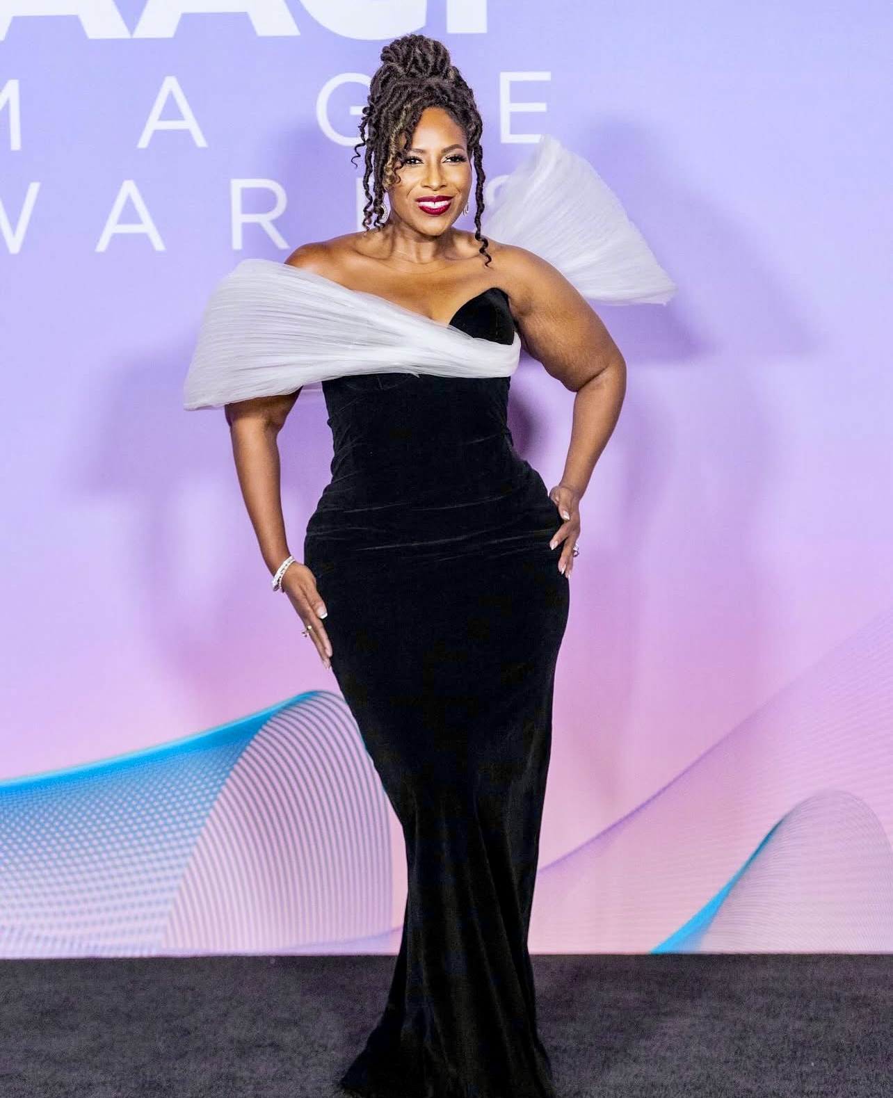 Woman in a black gown with white shoulder accents standing on stage with a purple background.