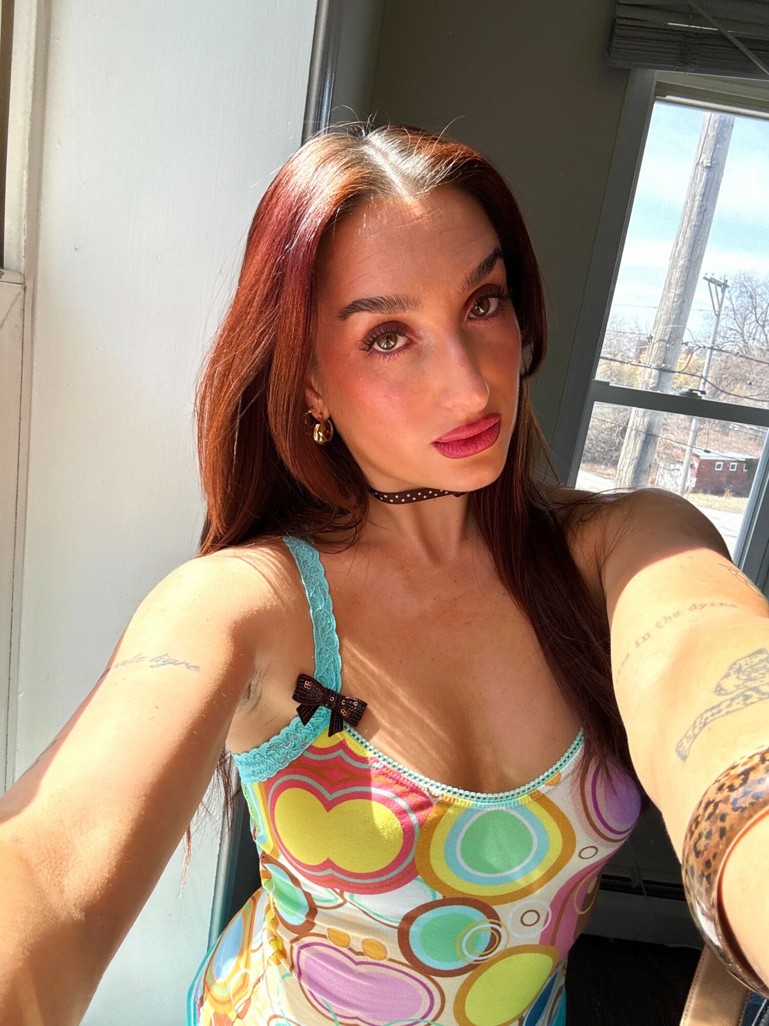 Young woman with long red hair taking a selfie indoors near a window, wearing colorful top and choker necklace.
