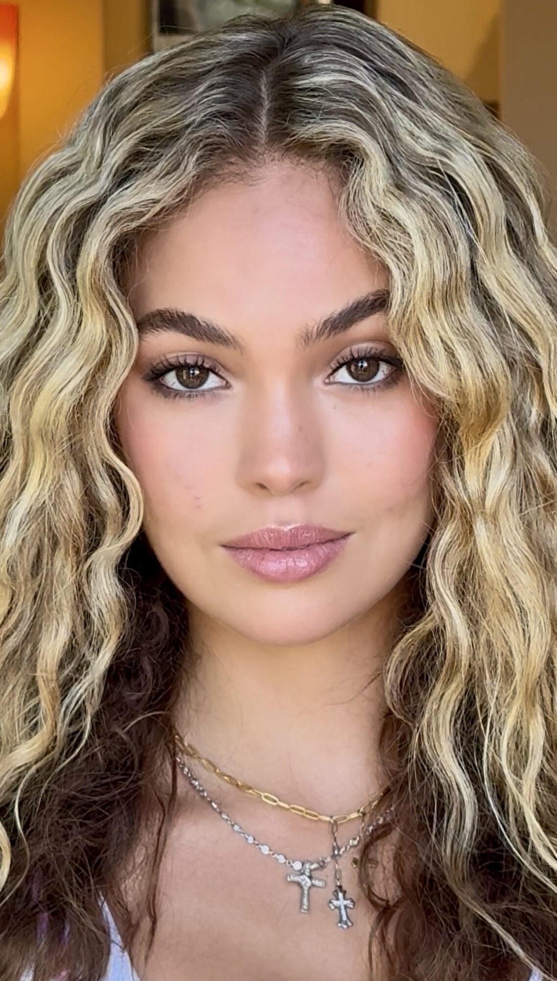Close-up of a woman with long, curly blonde hair and makeup, wearing layered necklaces, looking directly at the camera.
