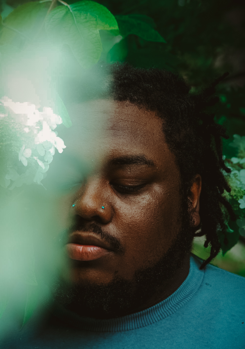 Close-up of a man with closed eyes, dark skin, and dreadlocks, surrounded by green foliage and a white flower.