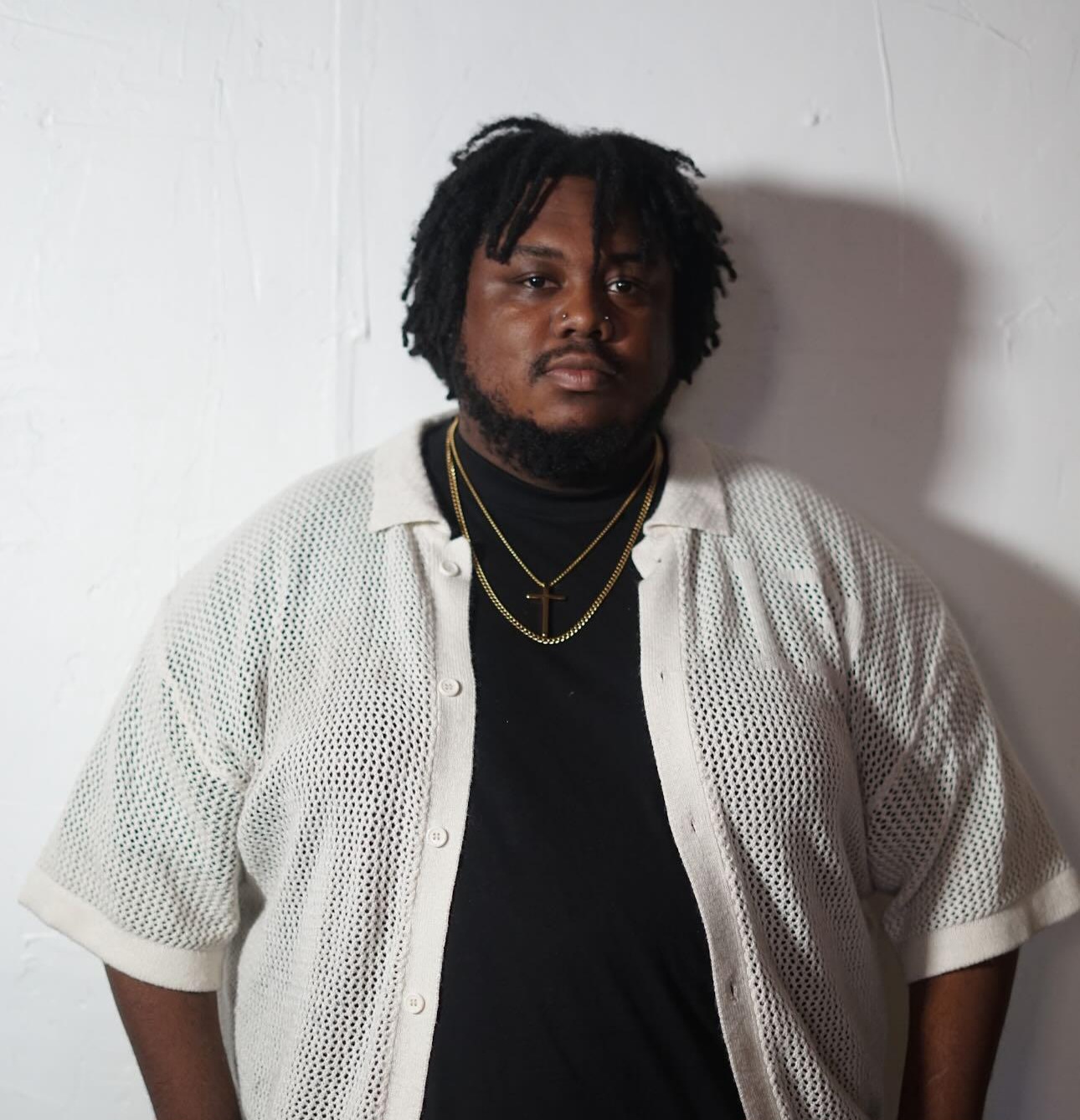 Person with dreadlocks wearing a white open shirt over a black shirt, standing against a white wall.