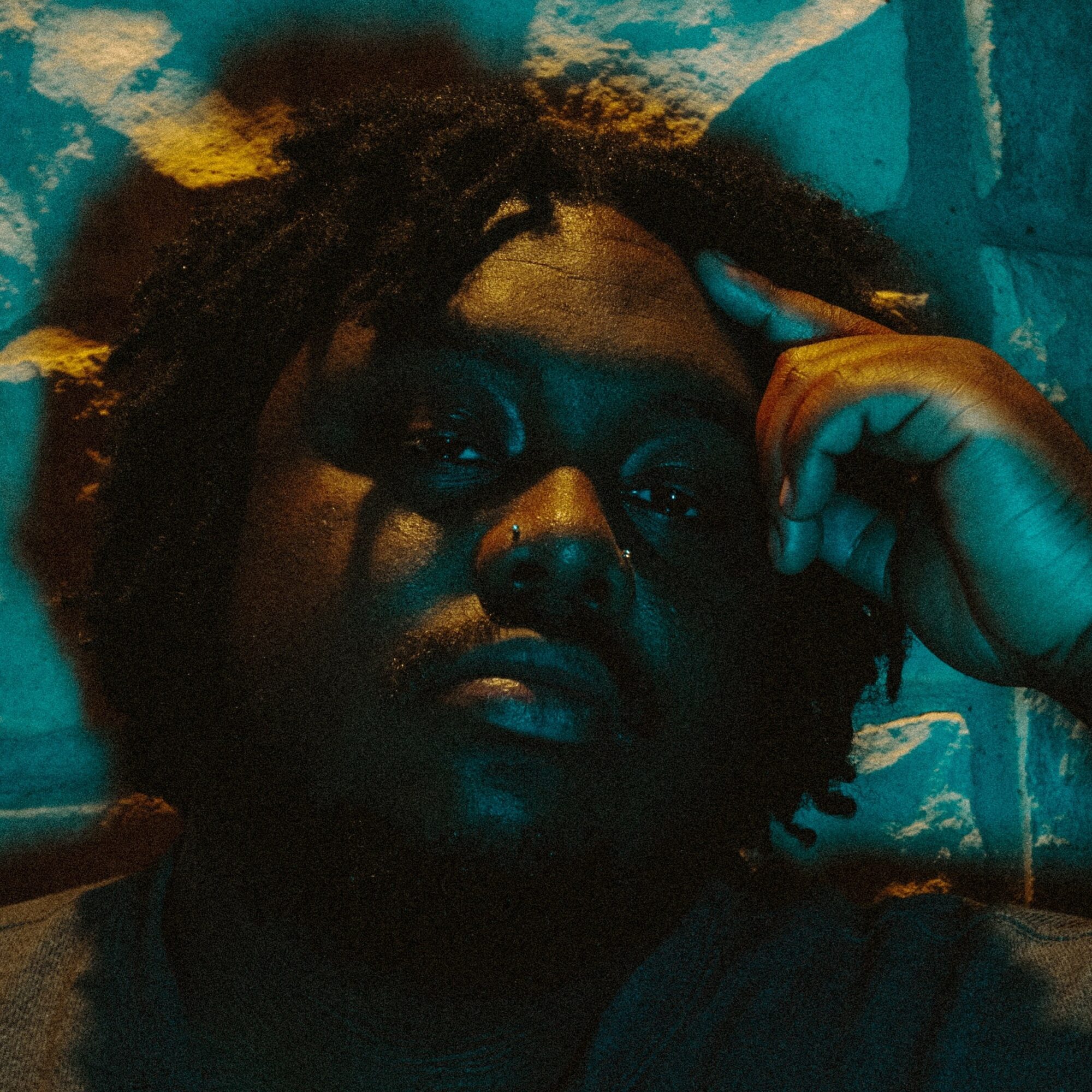 Person with dark skin and dreadlocks resting head on hand, in dim lighting, looking at camera.