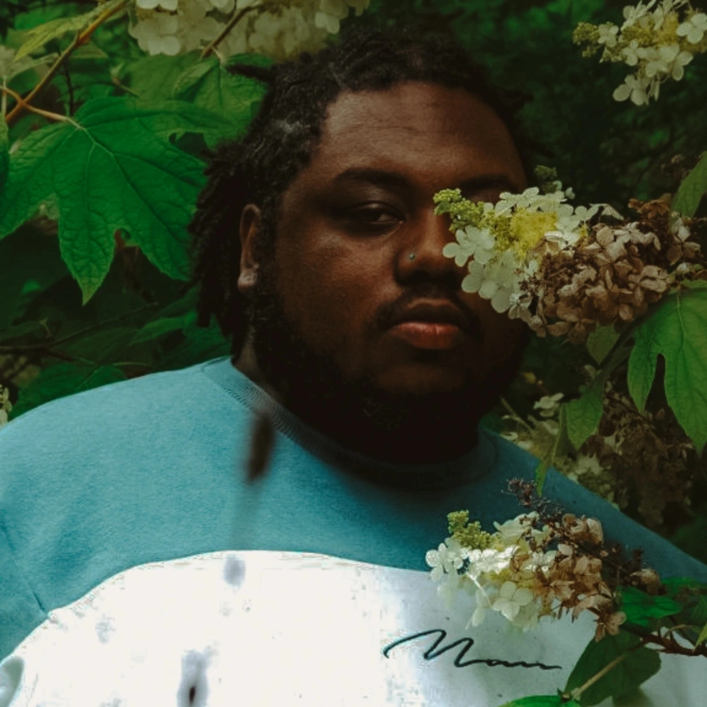 Person with dark hair and a serious expression among green leaves and white flowers.