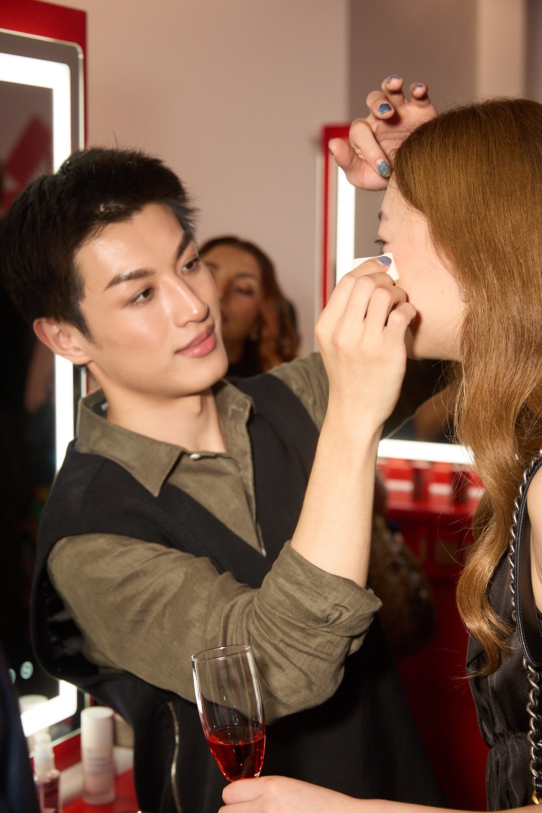 Makeup artist applies makeup to a woman's face in a beauty salon, with a glass of red wine on the counter.