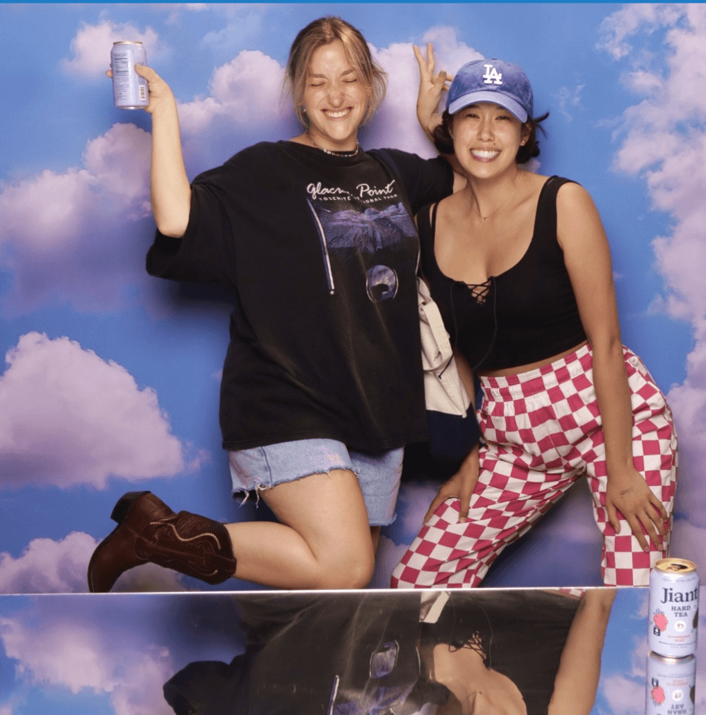 Two women smiling, one holding a drink, against a sky background. One wears a black shirt and denim shorts, the other a black top and checkered pants.
