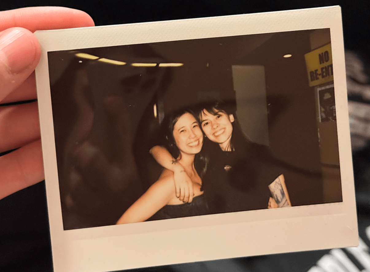 Two smiling women with arms around each other, posing indoors, with a dark background and some visible signage.