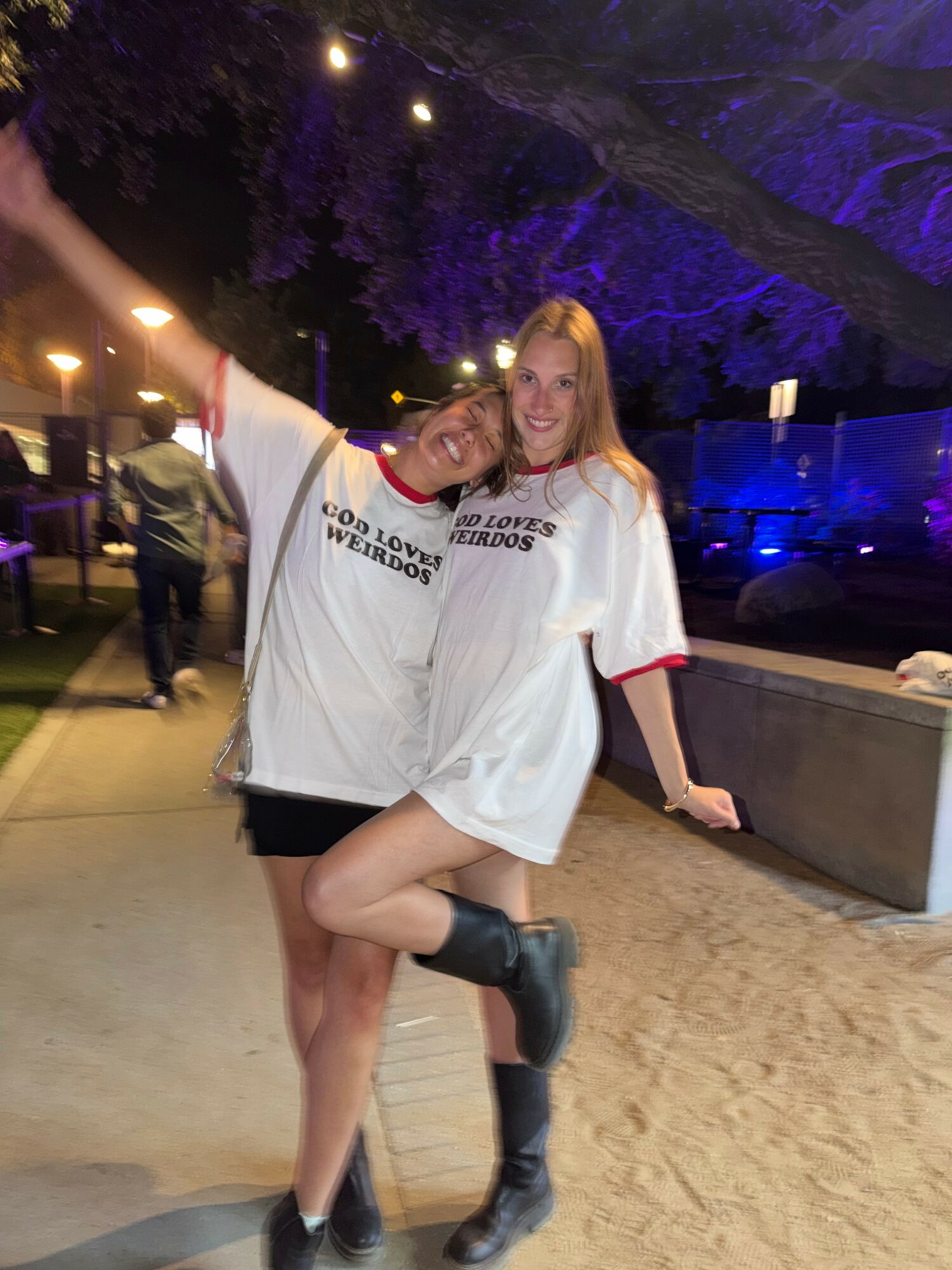 Two women standing outdoors at night, smiling, with trees and lights in the background.
