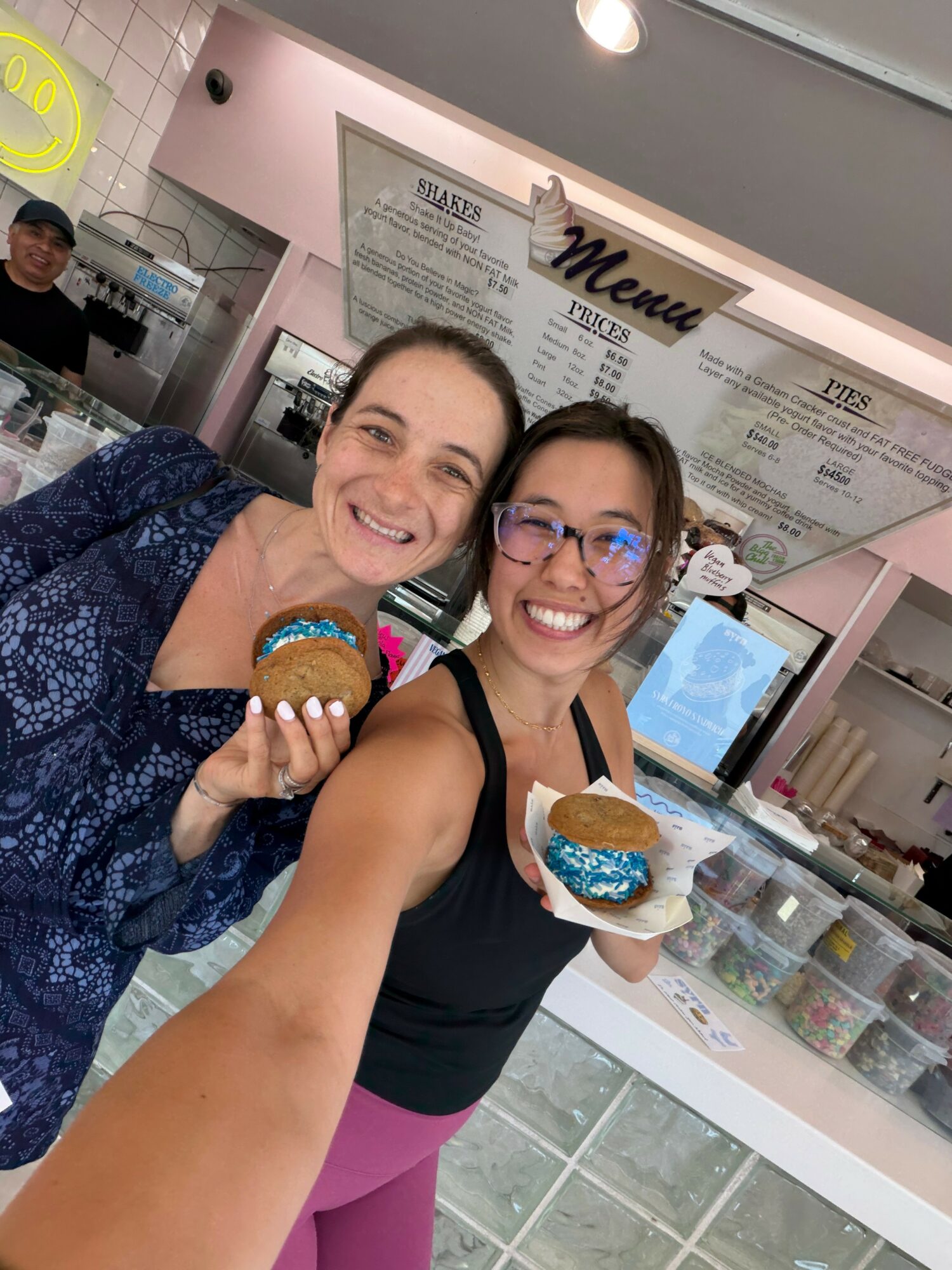 Two women smiling and holding cupcakes inside a bakery or cafe, with a menu board in the background.