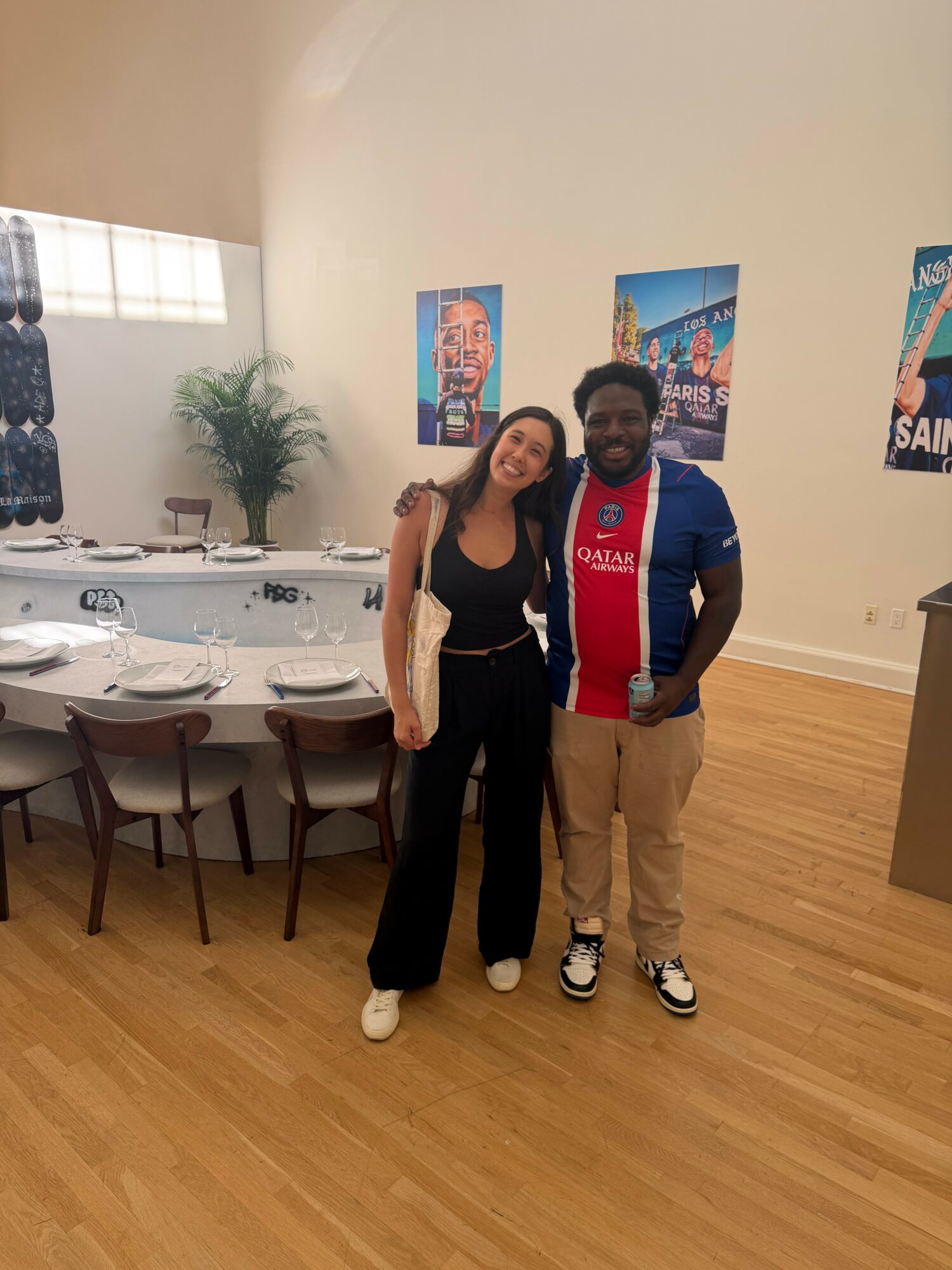 Two people standing together in a room with a table and chairs, posters on the wall, and a potted plant in the background.