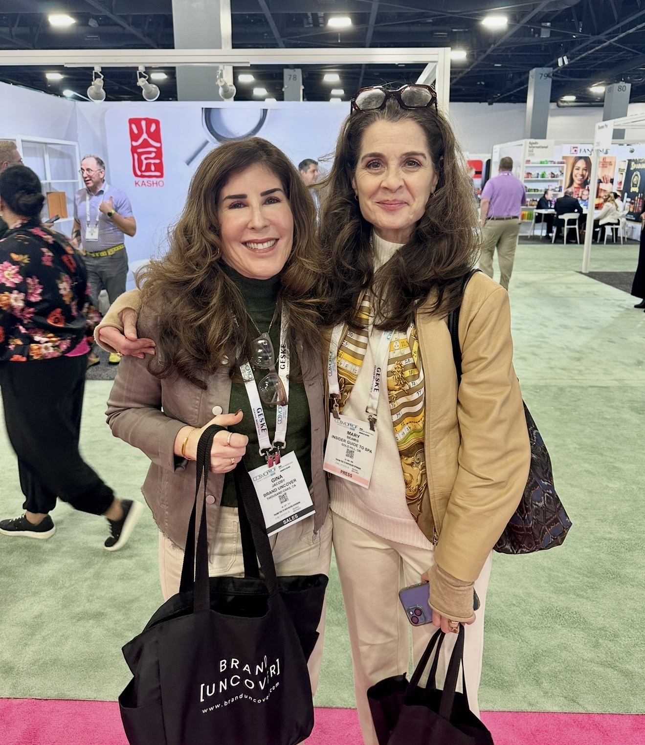 Two women smiling at an event, holding tote bags, with booths and people in the background.
