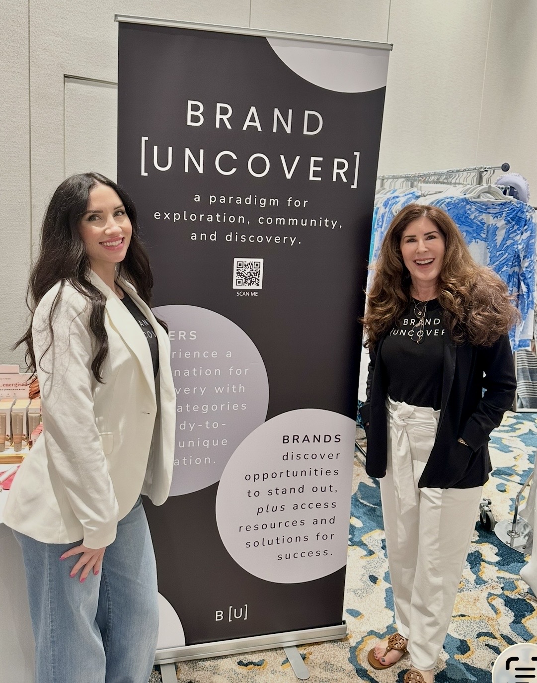 Two women standing next to a black and white vertical banner with text about branding and discovery, smiling indoors.