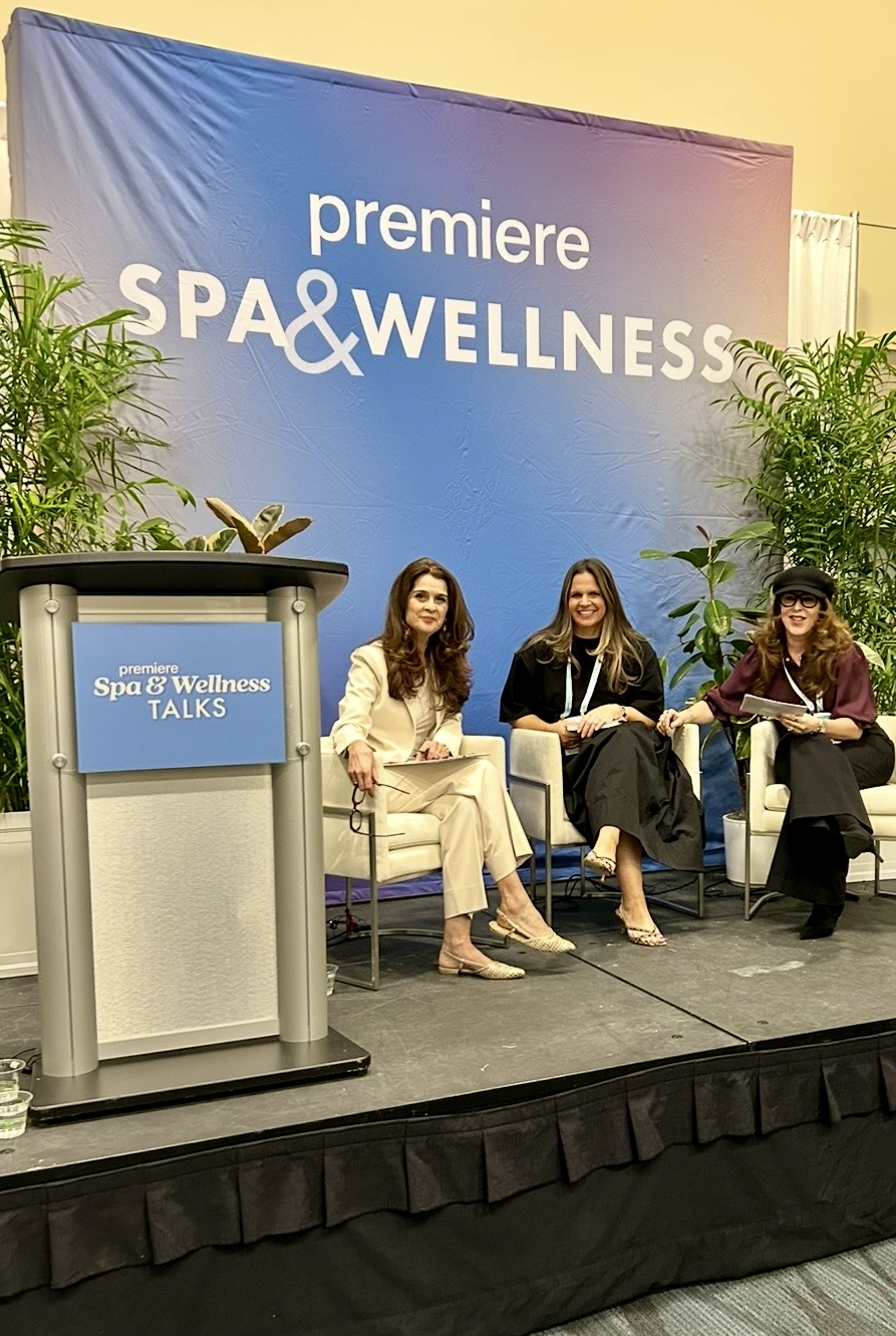Three women seated on stage at a spa and wellness event, with a blue backdrop and plants on either side.