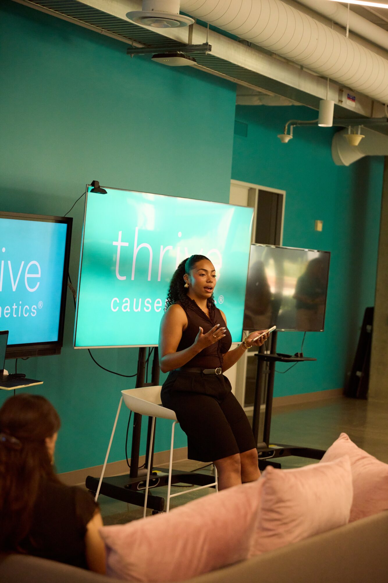 Woman standing and speaking in front of screens in a room with teal walls, audience seated nearby.