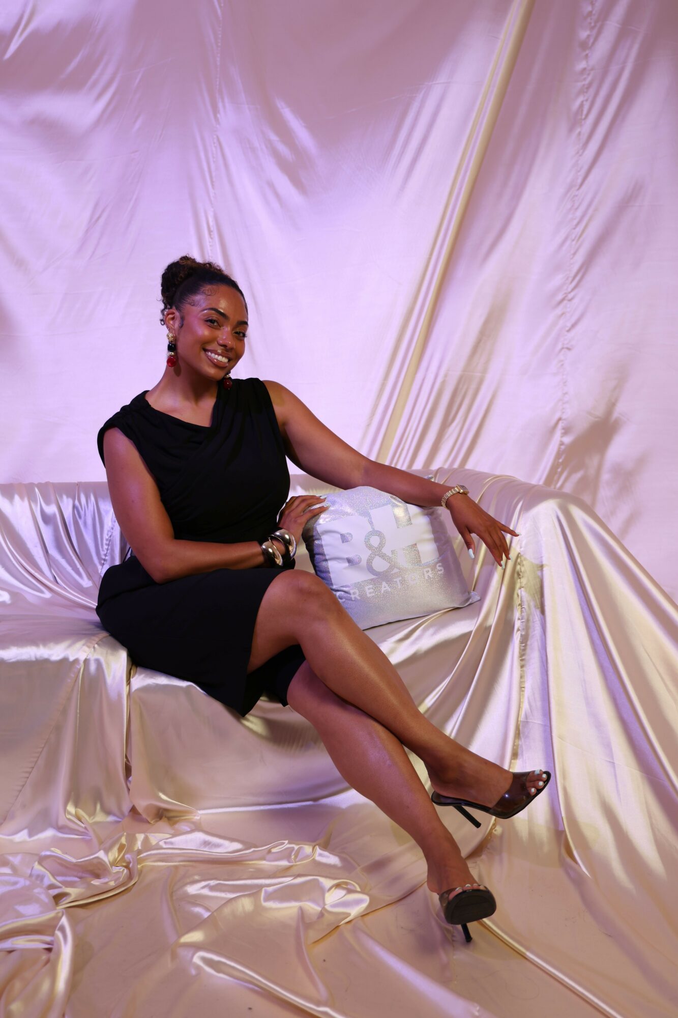 Woman in black dress lying on shiny, draped fabric, looking at camera, with one arm raised and legs crossed.