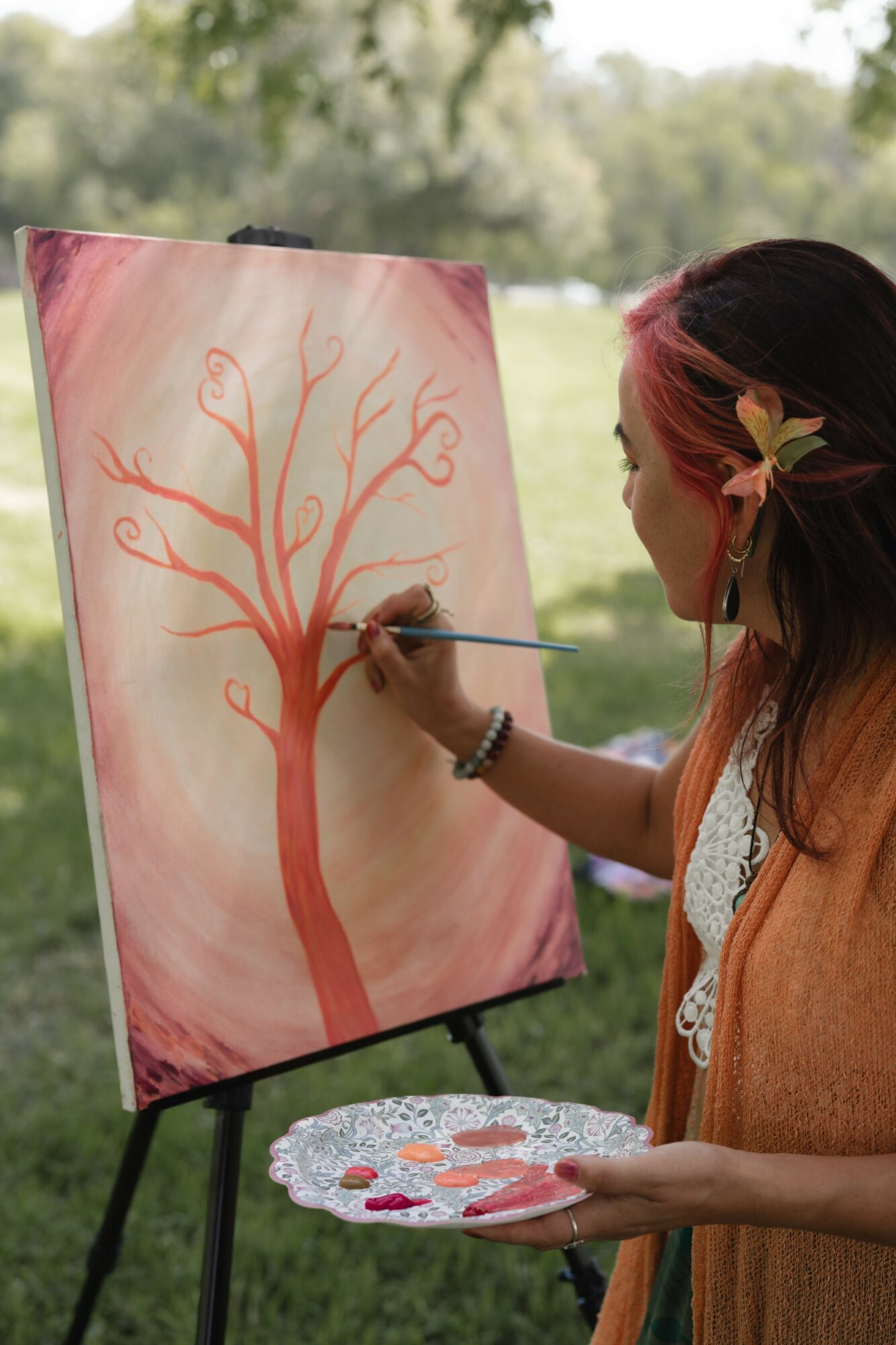 Woman painting a red tree on a canvas outdoors, holding a palette, with trees and grass in the background.
