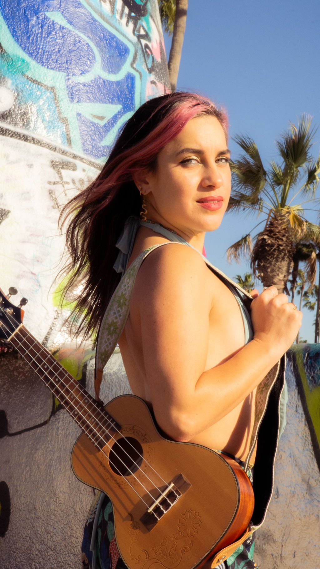 Young woman with pink hair holding a guitar, standing outdoors near a graffiti wall and palm trees.