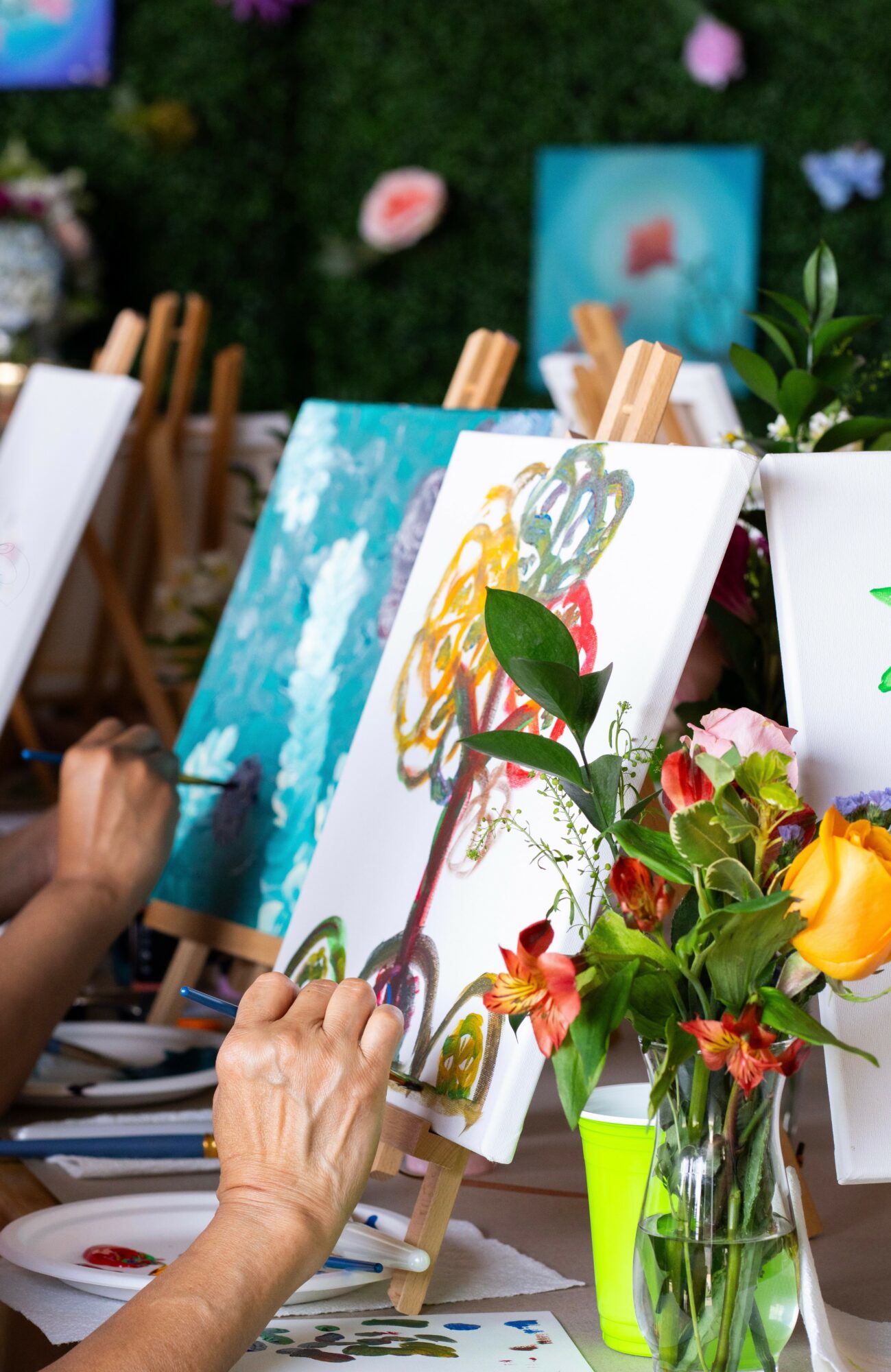 Person painting on canvas with colorful abstract design, surrounded by flowers and art supplies, outdoors.