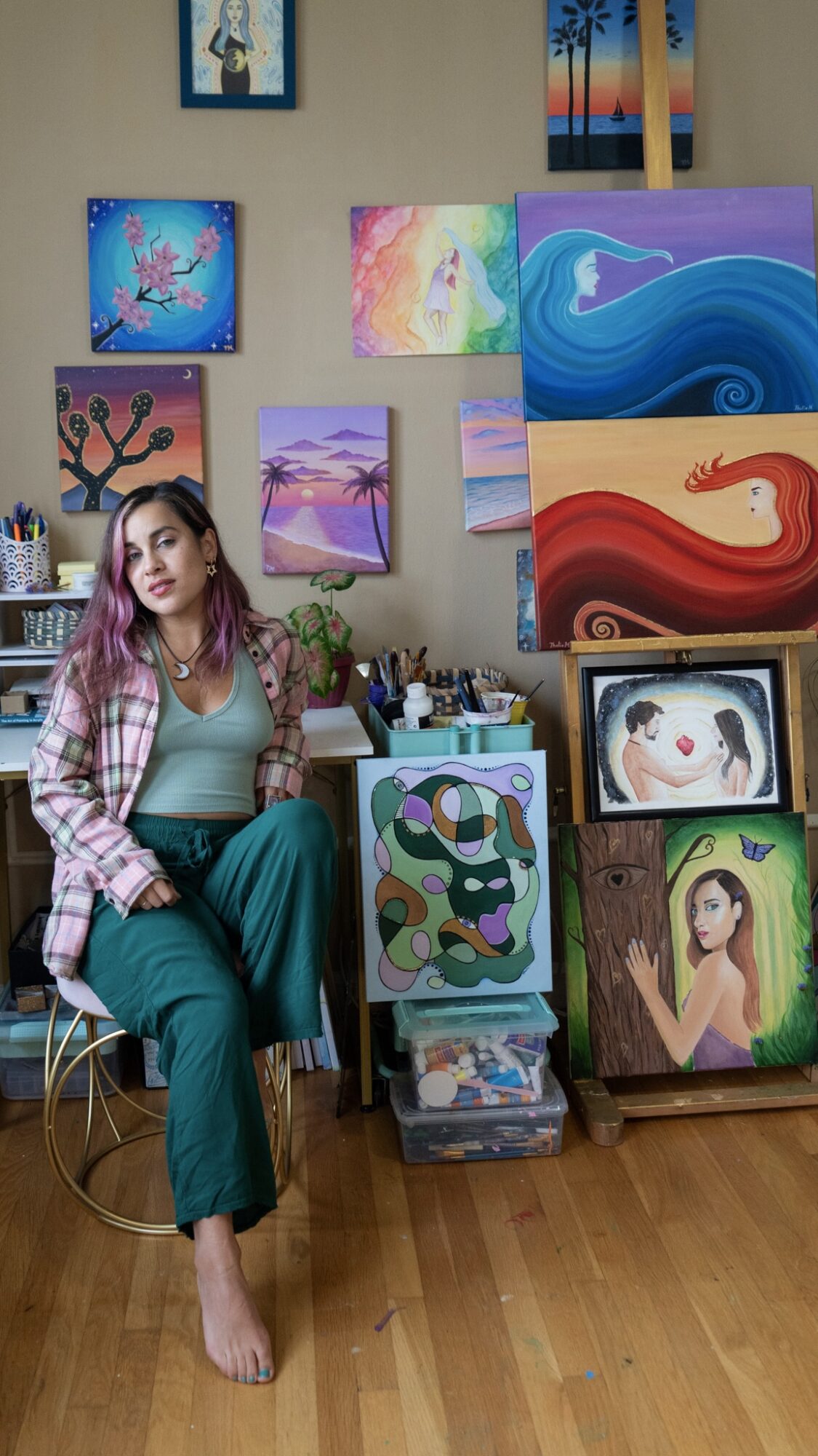 Young woman sitting on a chair in an art studio with colorful paintings on the wall and stacked on the floor.