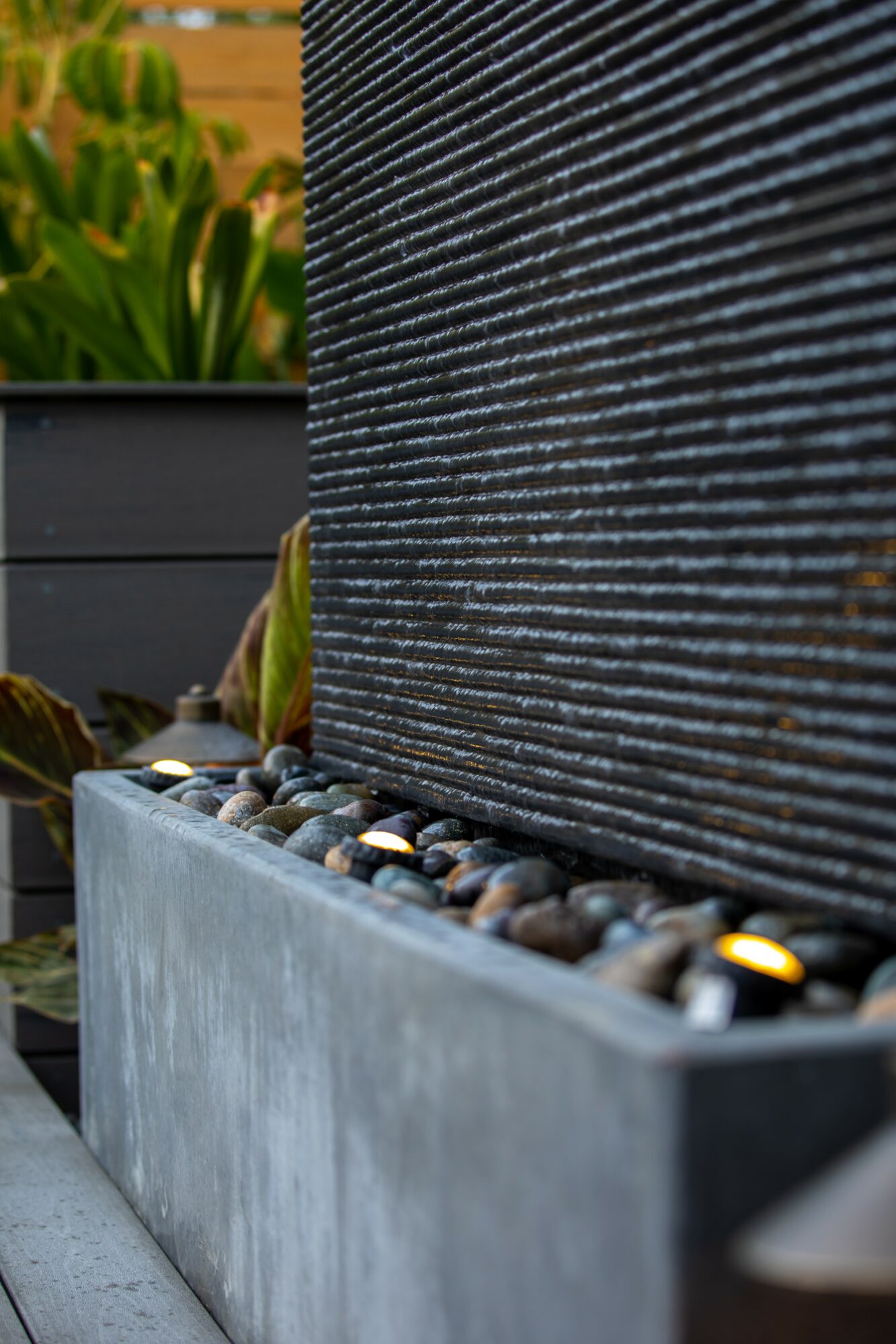 Close-up of a textured black wall with a concrete planter filled with small, smooth stones at the base.
