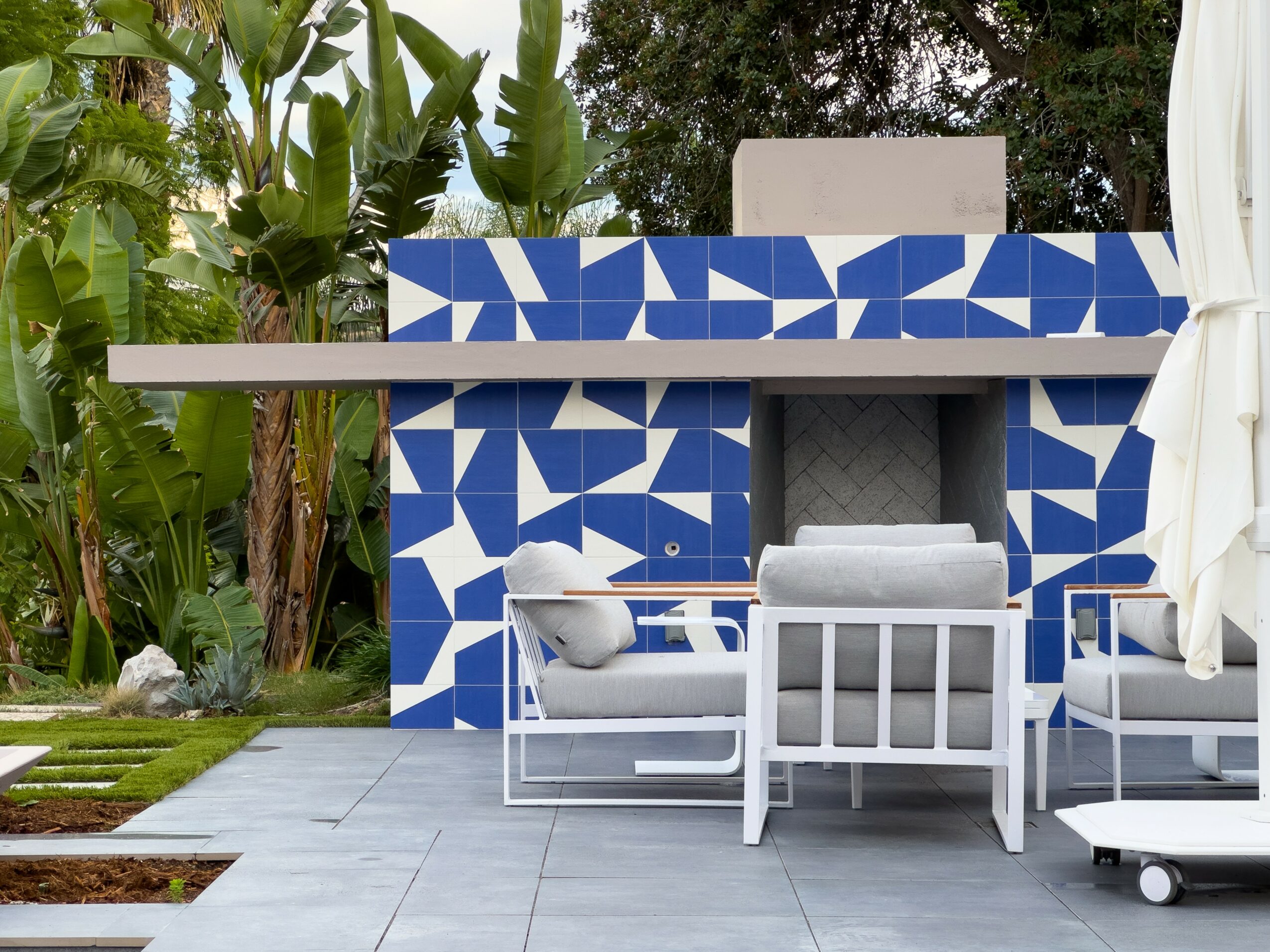 Outdoor patio with blue and white tiled wall, white chairs, and lush green plants in background.