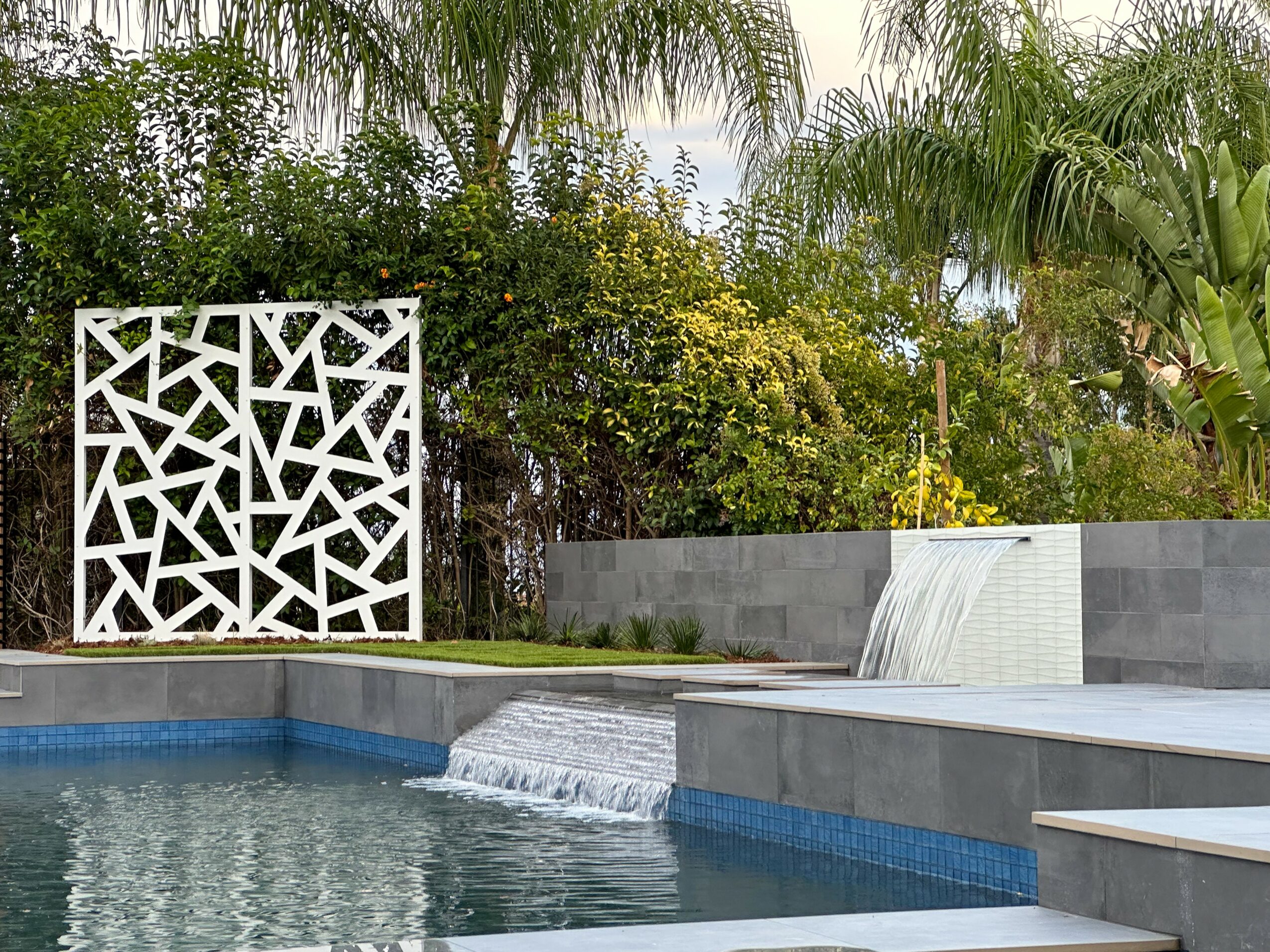 Swimming pool with waterfall feature, surrounded by greenery and modern gray stone structures.