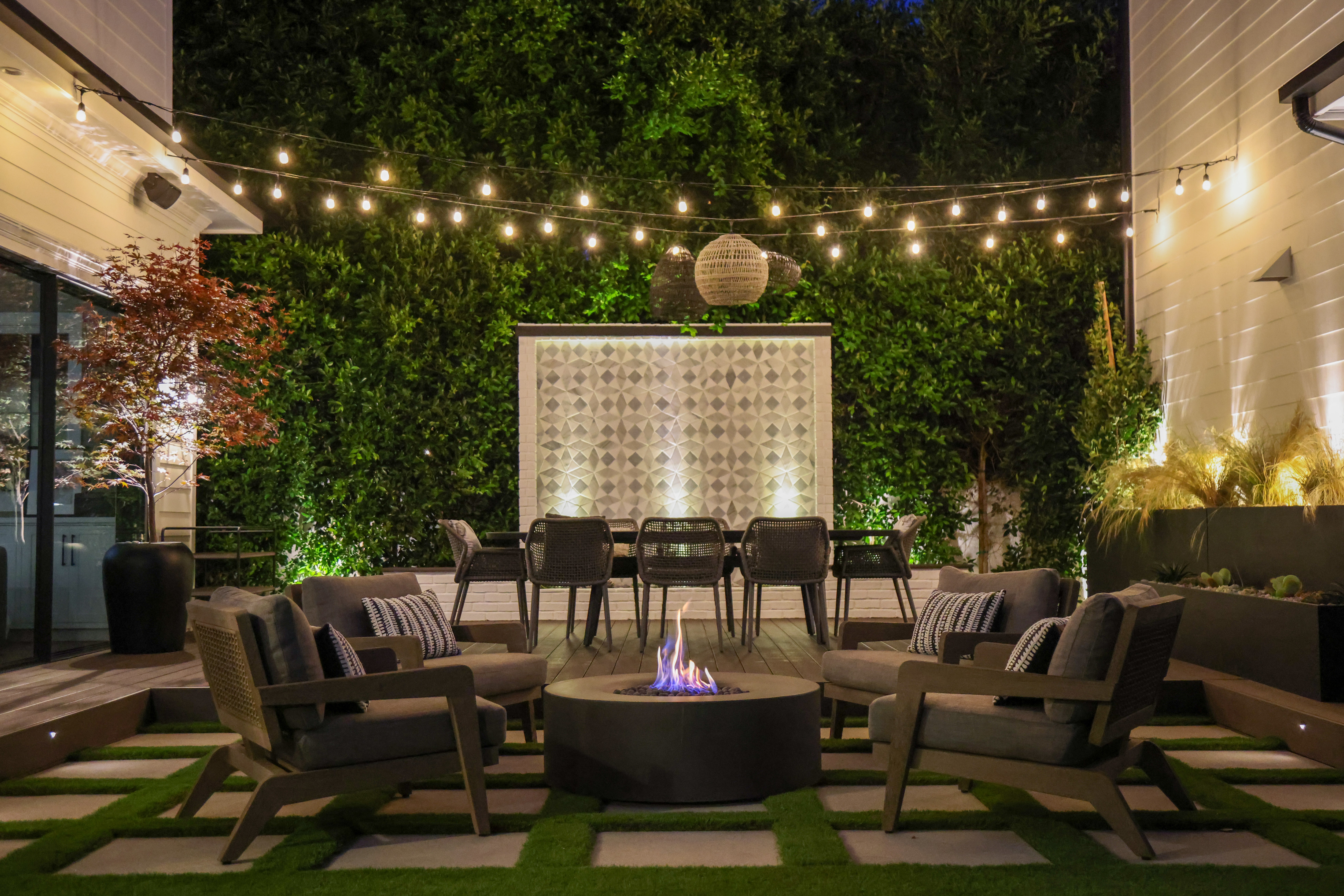 Outdoor patio with string lights, seating around fire pit, and greenery backdrop at night.
