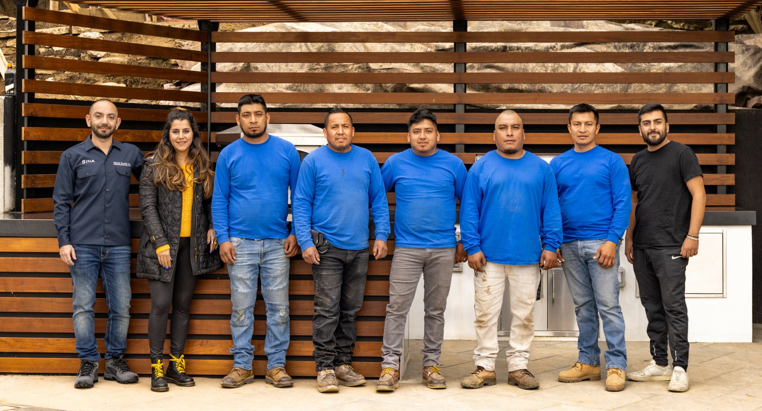 Group of seven people standing in front of a wooden fence, some wearing blue shirts, one woman in a black jacket, and one man in black shirt, all smiling.