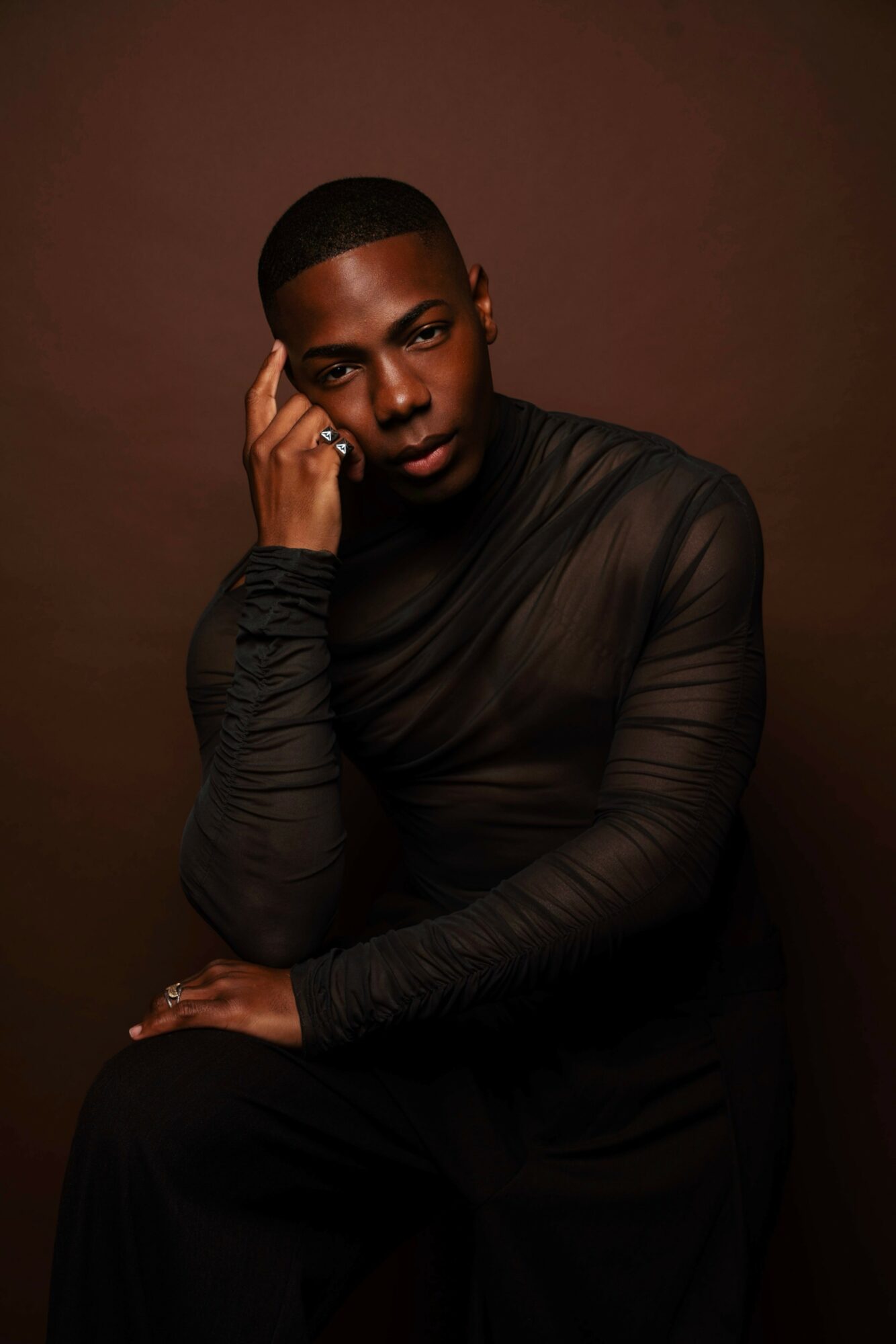 Portrait of a person with short hair, wearing a sheer black top, posing with hand near face against a brown background.