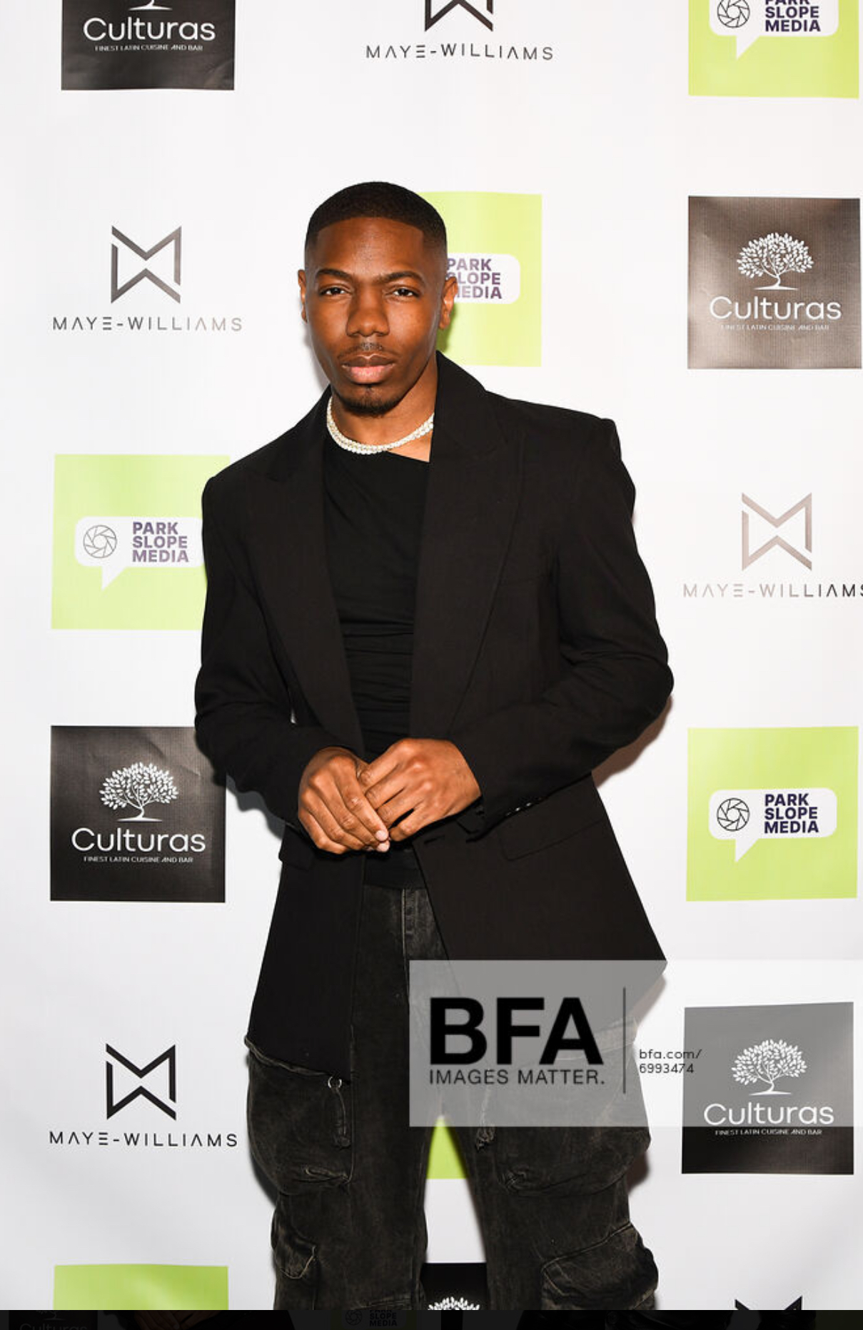 Young man in black blazer and jeans standing in front of a step-and-repeat backdrop with logos.