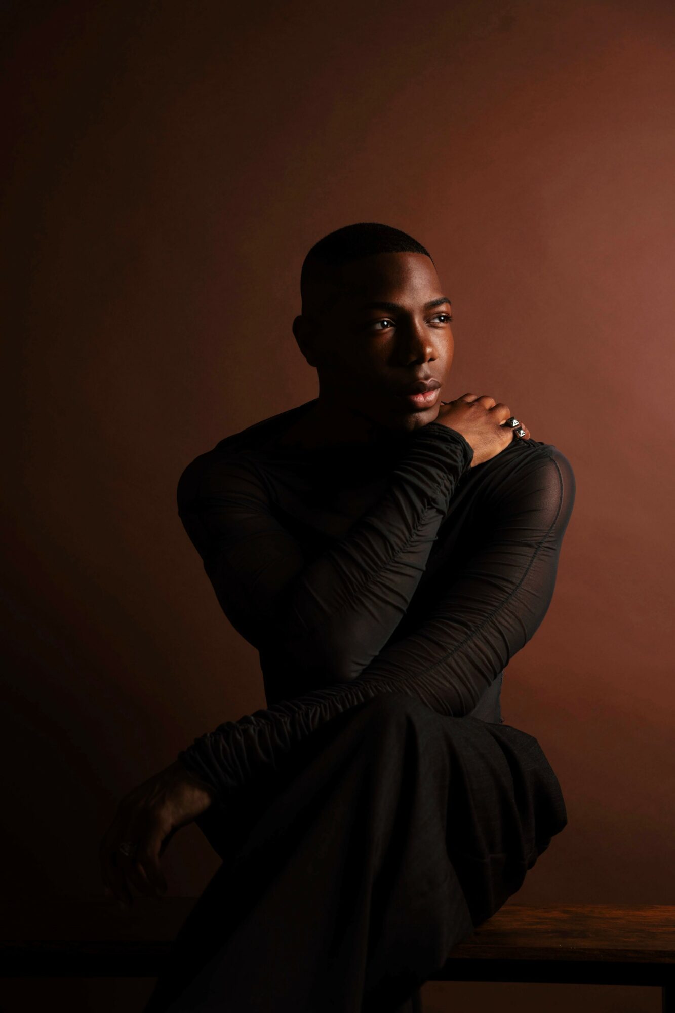 Portrait of a person with short hair, wearing a black long-sleeve top, sitting with arms crossed, against a brown background.