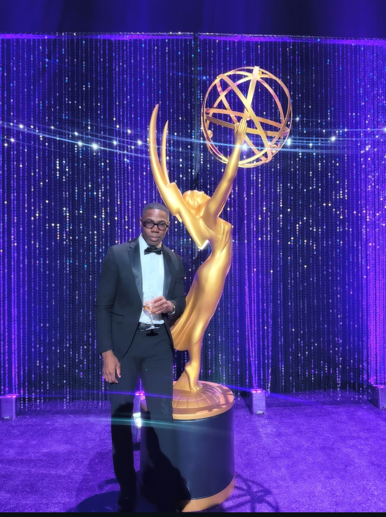Man in tuxedo standing next to a large golden Emmy statue with a purple curtain background.
