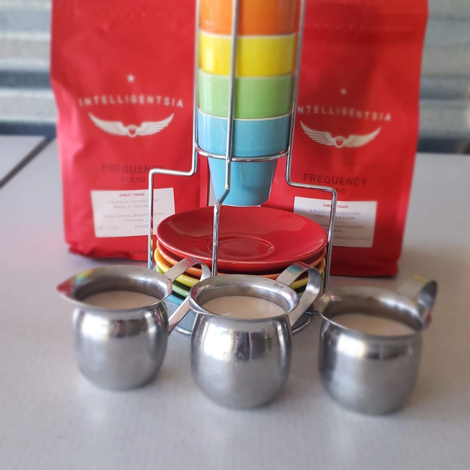 Three metal measuring cups in front of colorful rainbow stacking toy and red coffee bags with white text and logo.