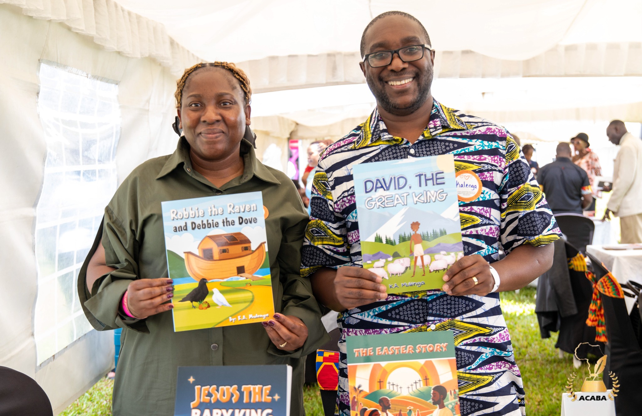 Two people holding children's books, smiling, indoors with other people in the background.