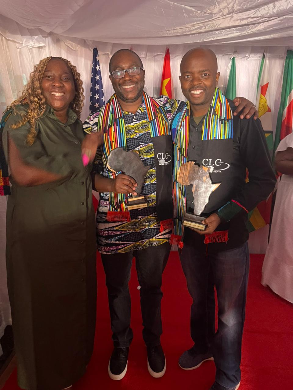 Three people standing together, smiling, with flags in the background, one holding a trophy, and another holding a plaque.