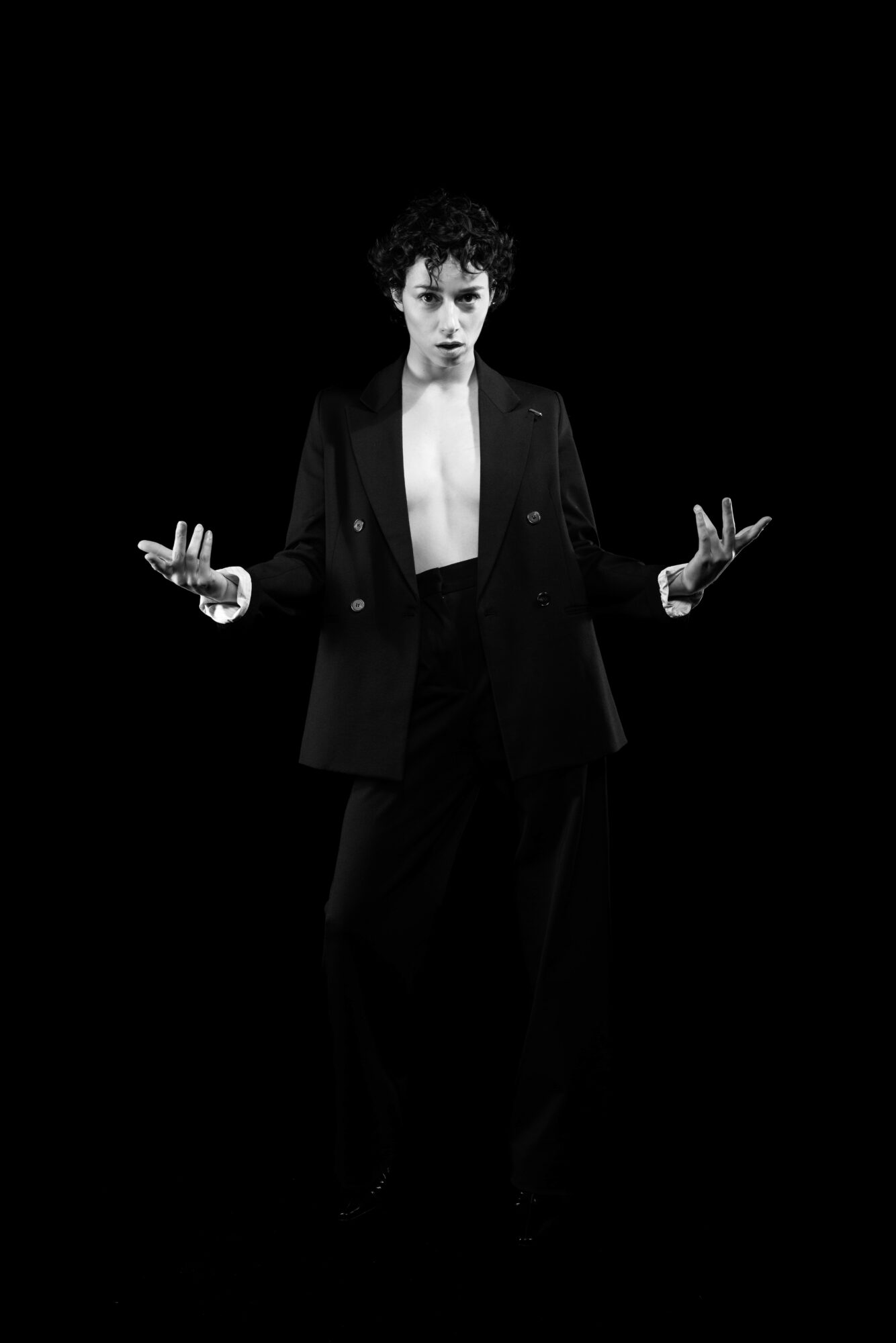 Person with short curly hair wearing a blazer, standing with hands raised, against a black background.