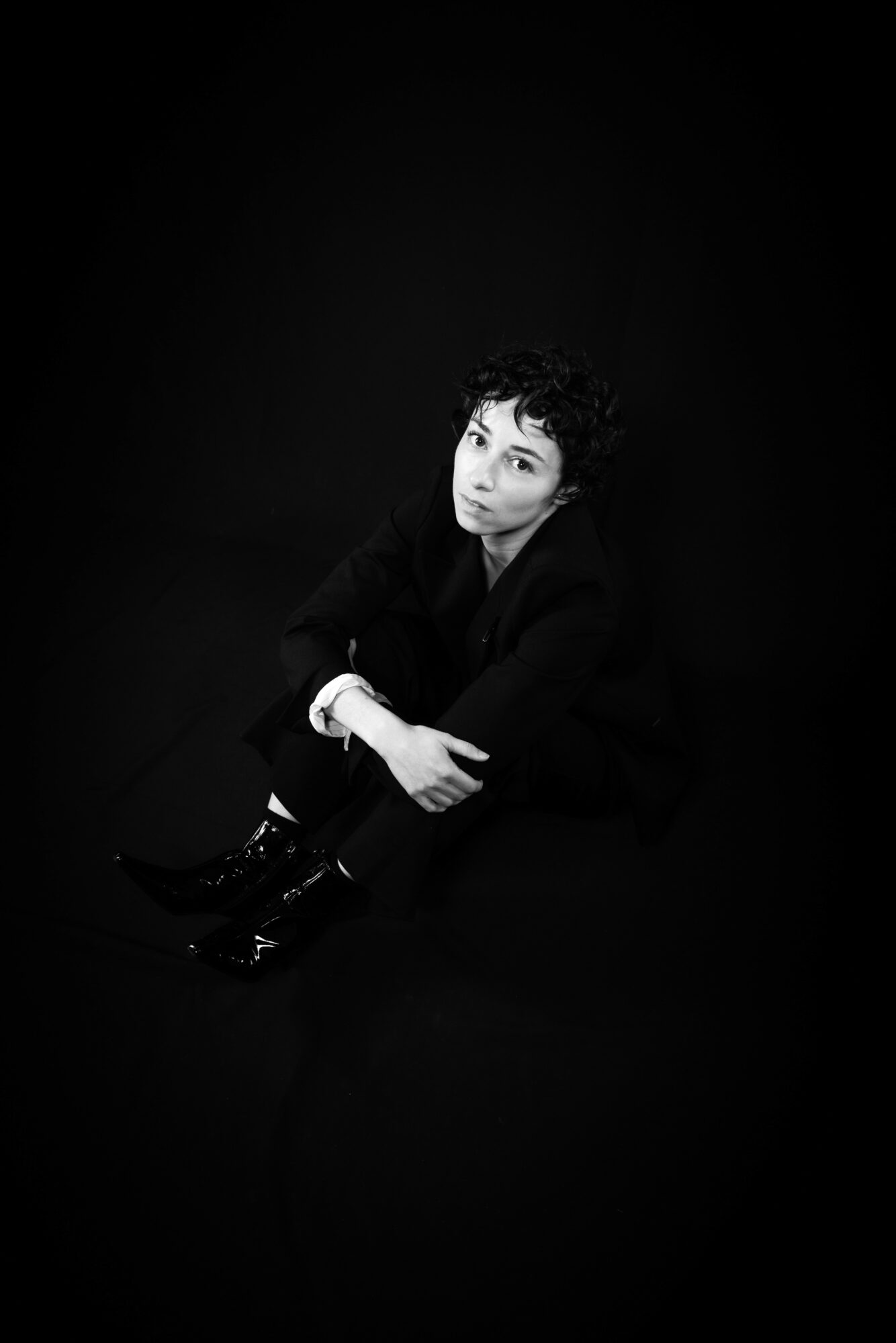 Person sitting with crossed arms, looking at camera, in dark clothing against a black background.