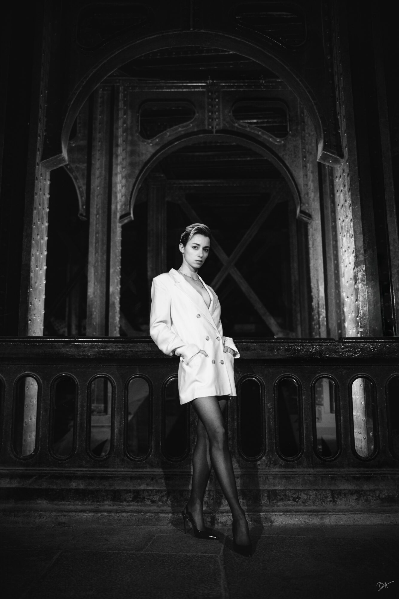 Woman in a light coat and dark tights standing on a balcony in an ornate, historic building interior.