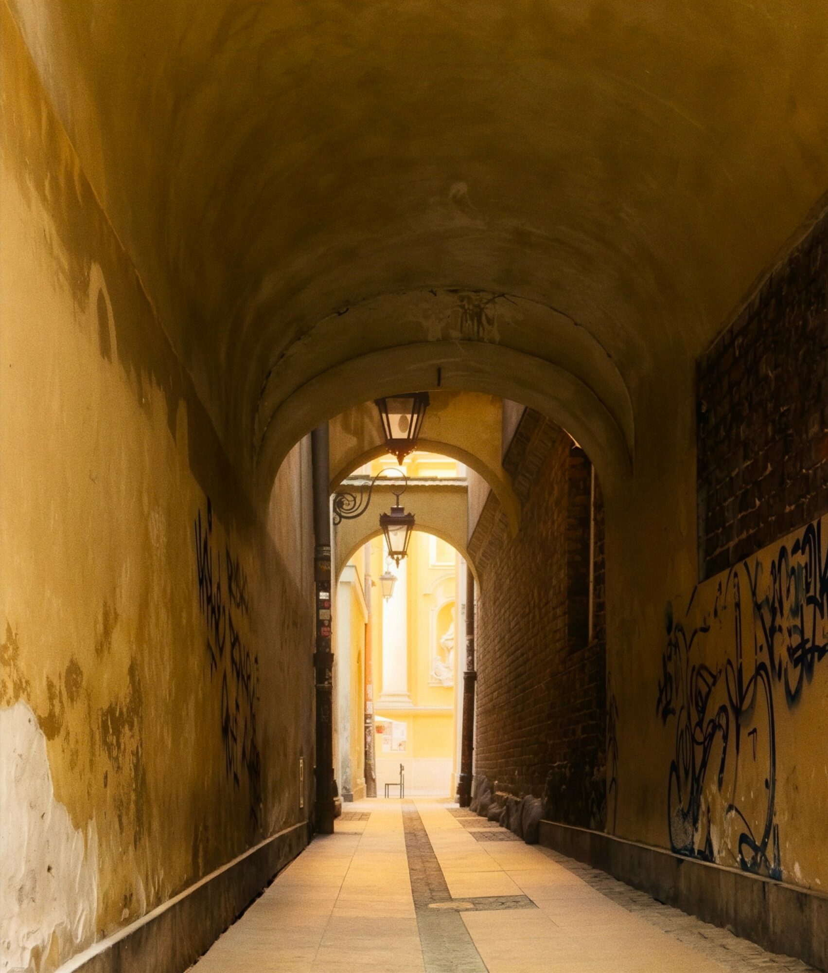 Narrow alleyway with arched ceiling, graffiti on right wall, and light at the end, leading to a bright street scene.
