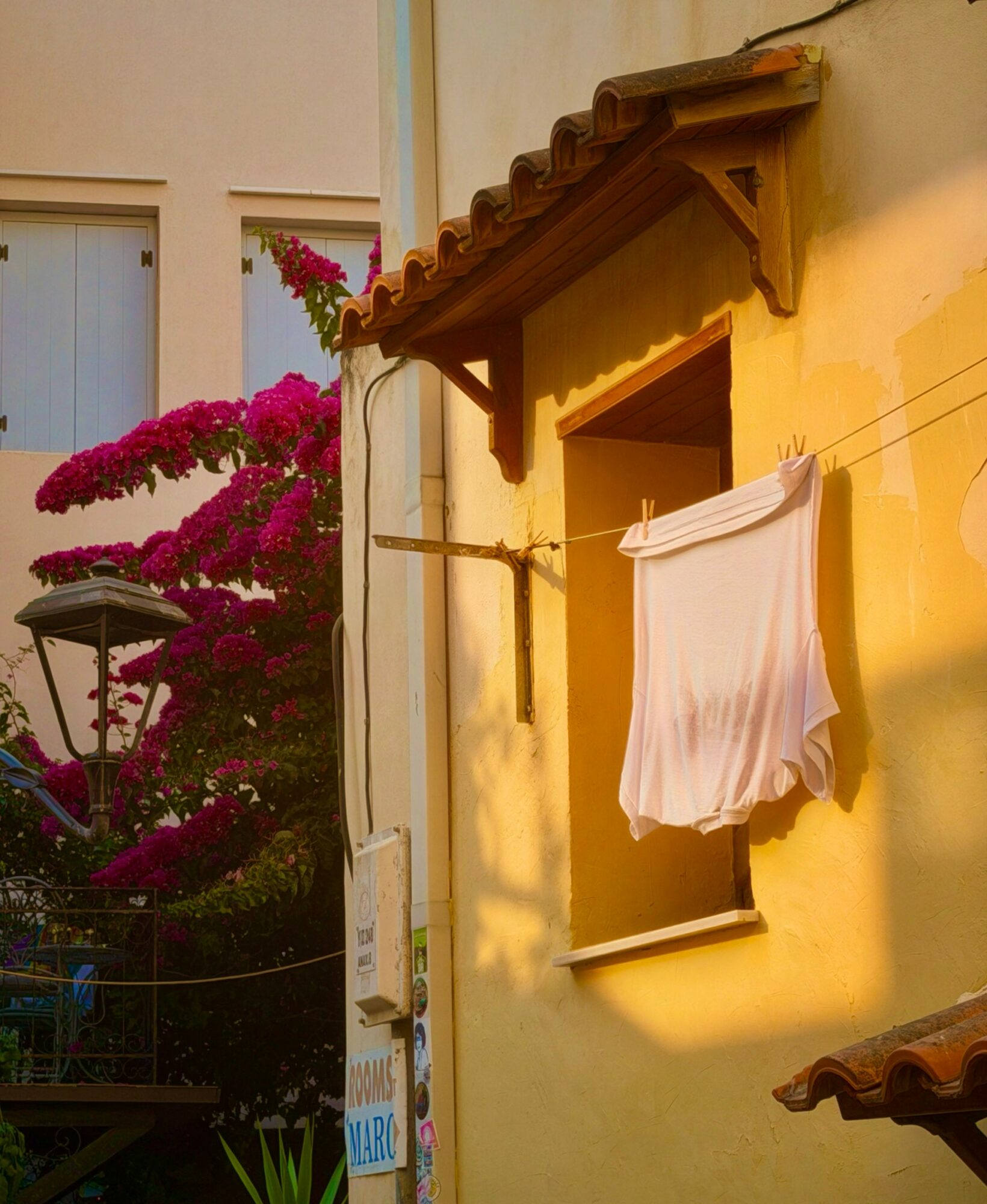 Sunlight illuminates a yellow wall with a window and a white cloth hanging outside, with pink flowering bushes nearby.