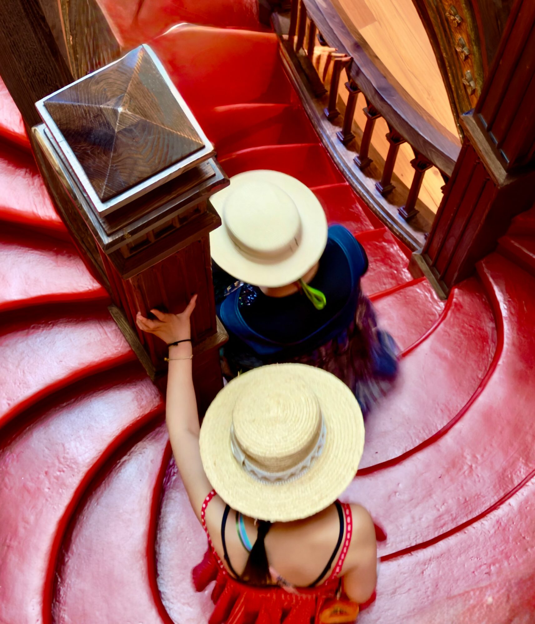 Two women with wide-brimmed hats sitting on a red curved staircase, viewed from above, with a wooden railing nearby.